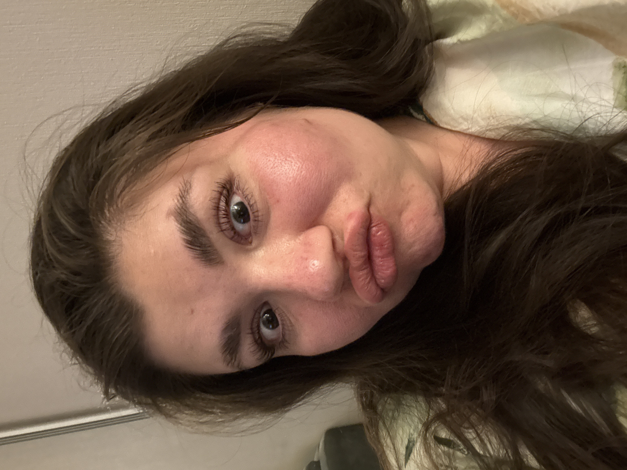 A close-up, angled self-portrait of a Caucasian woman with long, dark brown hair. She is looking at the camera with a slight pouting expression. Her eyes are wide and her eyebrows are thick and dark. Her skin appears slightly flushed. She is lying on a patterned bedding. There is no indication this image was taken in Votkinsk, Russia.