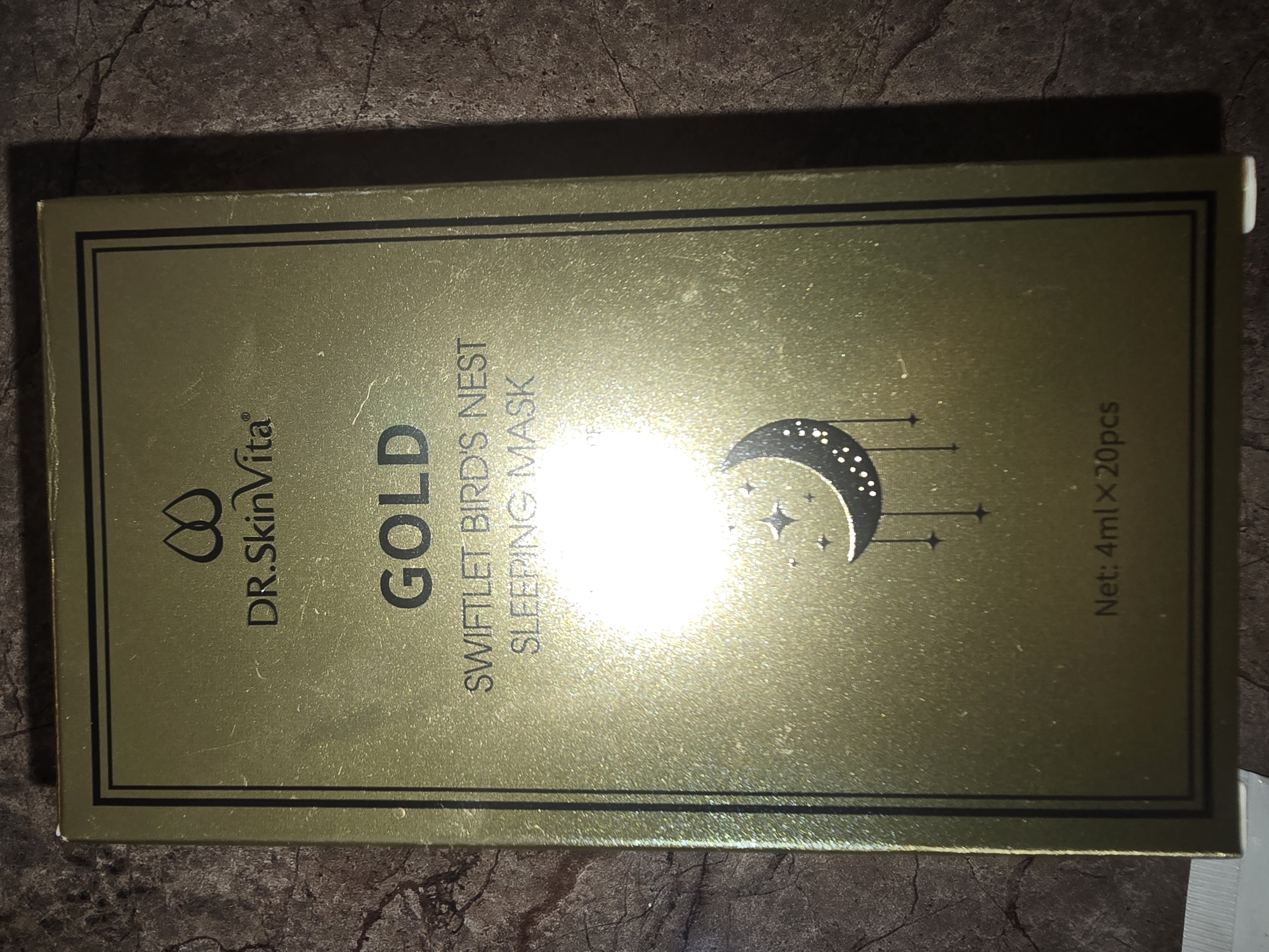 The image displays the front of a gold-colored cardboard box for a skincare product. The box has a dark border and contains text and an illustration. At the top, the text "Net: 4ml x 20pcs" is visible. Below this, an illustration of a crescent moon with stars is present. Further down, the words "GOLD" are prominently displayed. Beneath "GOLD", smaller text reads "SWIFTLET BIRD'S NEST SLEEPING MASK". At the bottom of the box, "DR.SKINVita" is written, accompanied by a symbol resembling an infinity loop. The box is placed on a dark, textured surface, possibly a table or countertop. There are no people or animals present in the image. The lighting suggests the photo was taken indoors, with a bright light source creating a glare on the box. The location of Ulan-Ude, Russia, is not depicted or implied by the image content.
