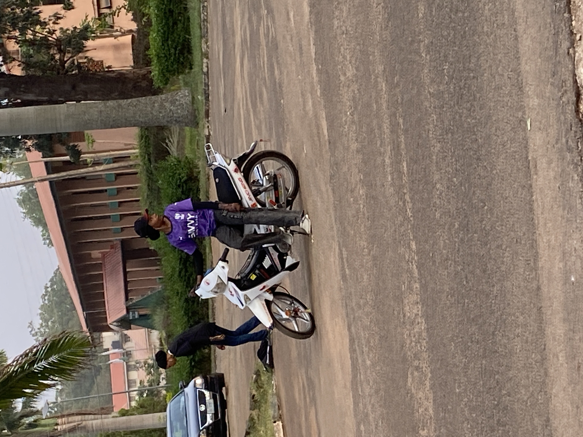 An outdoor, eye-level, full shot depicts two men and their motorcycles parked on the side of a paved road in Vom, Nigeria, during the daytime. The image is oriented vertically (rotated 90 degrees clockwise).
In the center, a man wearing a purple t-shirt with white text that reads "SAVVY", dark pants, and a black cap stands leaning against a white and black motorcycle. He looks towards the viewer. To his right and slightly behind him, another man in a dark long-sleeved shirt and blue jeans is bent over near a second white and black motorcycle, which is partially obscured. A dark-colored car, possibly a sedan or SUV, is parked further down the road on the far right, with its front end and headlights visible.
The setting features a wide, grey-brown paved road in the foreground and extending into the background on the right. Lush green vegetation, including bushes and tall trees with slender trunks, lines the left side of the road. In the background, several buildings with brown roofs and light-colored walls are visible, suggesting an institutional or residential area. The sky appears bright but slightly overcast, indicating a clear day. No strong emotions are discernible from the individuals.
