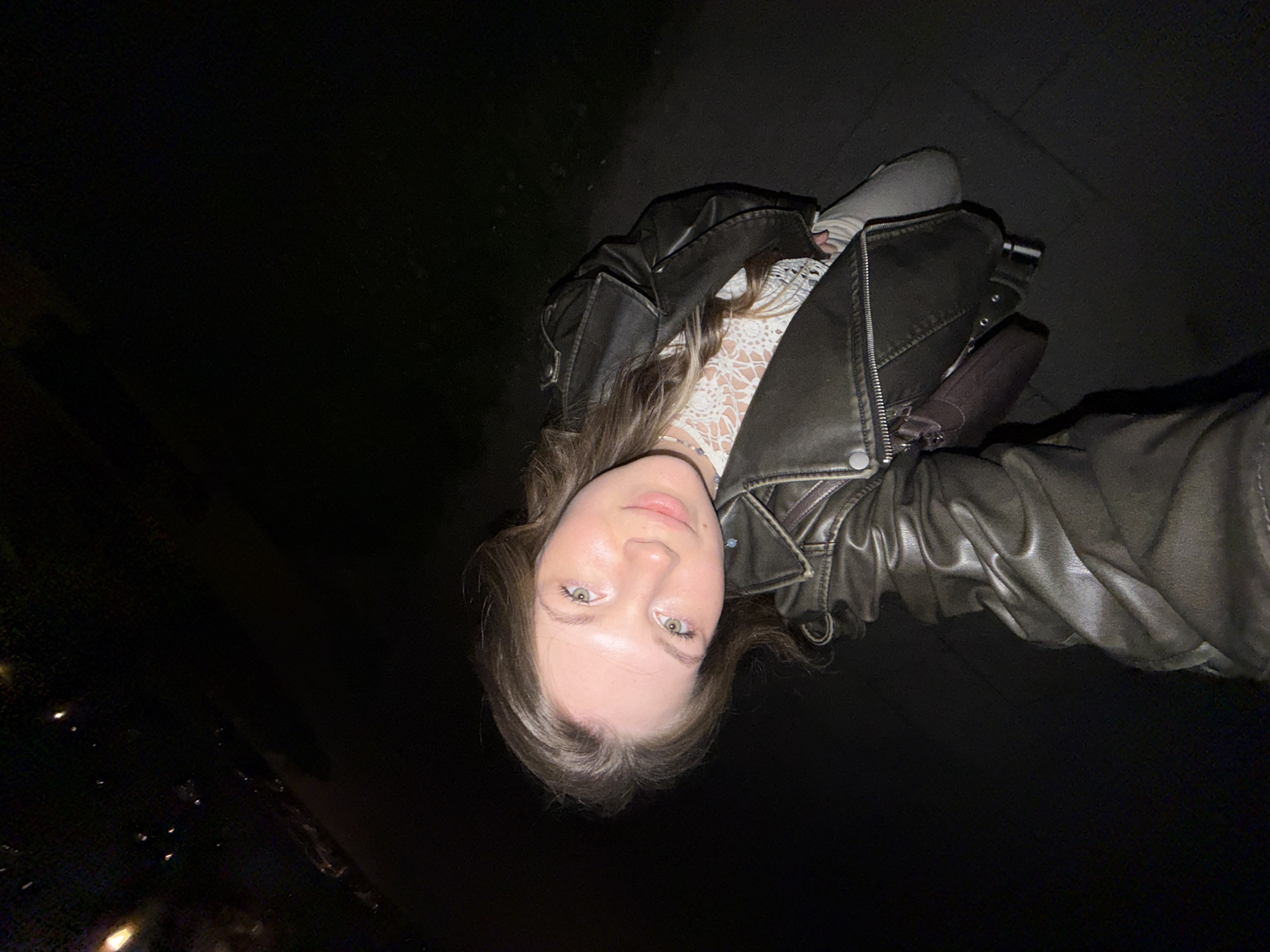 A young woman is posing for a selfie at night in Belgrade, Serbia. She is wearing a brown leather jacket and a white lace top. The image is taken at night, and the background is dark. In the lower left of the image are city lights.
