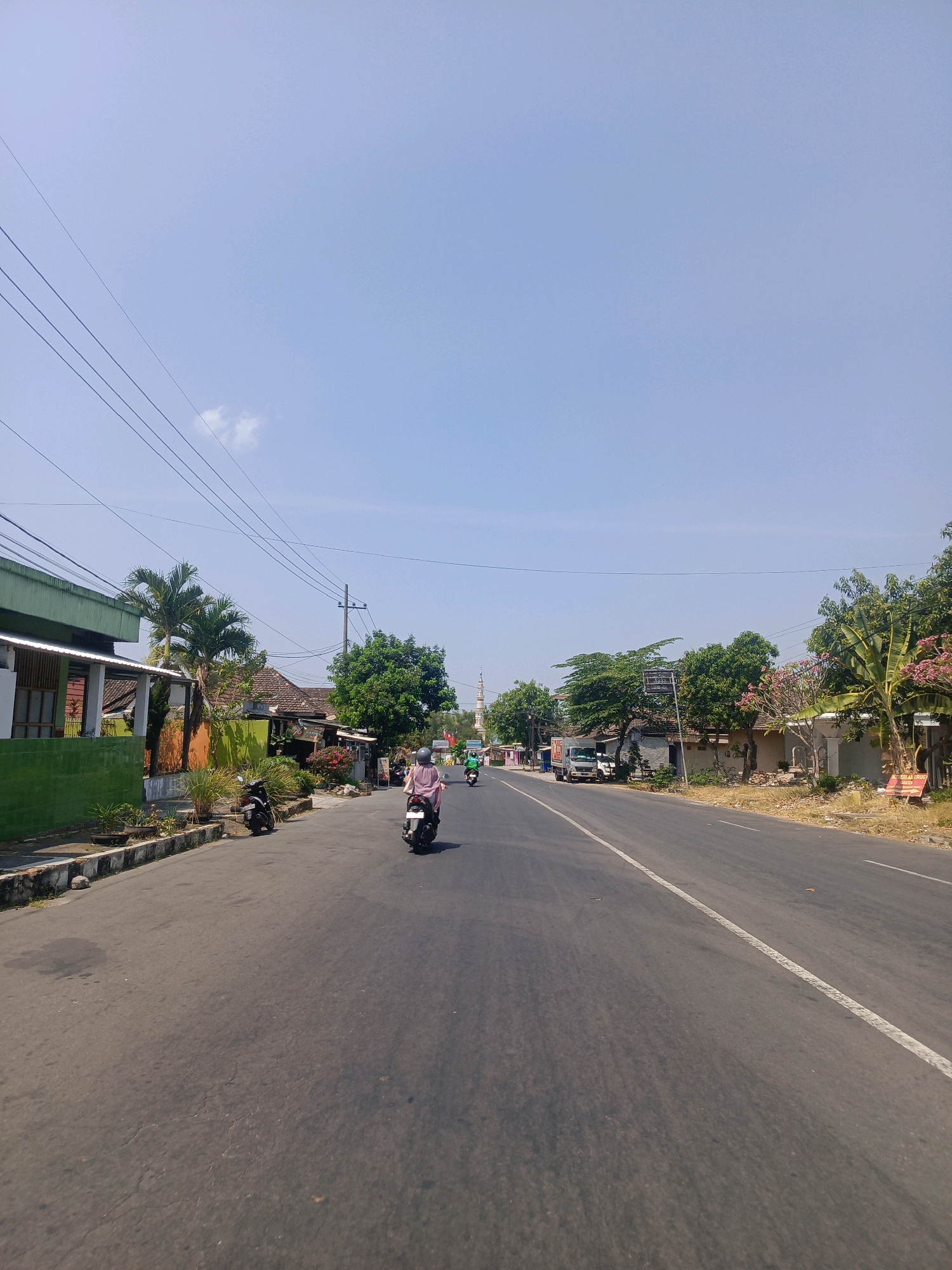 The image shows a sunny day on a paved road in Milangasri, Indonesia. The road is lined with low-rise buildings, some with green facades, and lush vegetation including palm trees and flowering bushes. Several motorcycles are visible on the road. In the foreground, a person wearing a pink outfit rides a scooter. Further down the road, other motorcycles and a truck can be seen. A tall, slender structure resembling a minaret stands in the distance on the right side of the road, suggesting a mosque in the vicinity. The sky is clear blue with a few wispy clouds. Power lines crisscross the upper portion of the frame. The overall scene depicts a typical streetscape in a tropical, possibly rural or semi-urban, Indonesian setting.