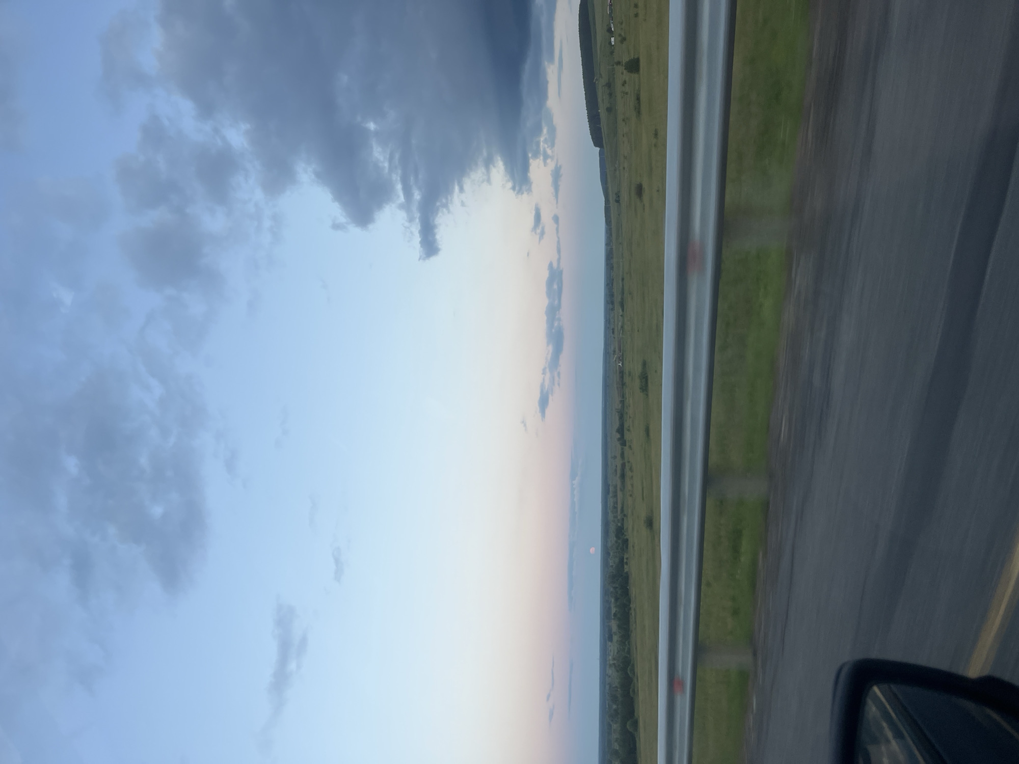 The image captures a wide landscape view of a rural area, possibly near Tarasovo, Russia, during what appears to be dusk or dawn. The sky is a gradient of light blue and pale pink, with scattered clouds, some of which are dark and ominous. In the distance, a faint, reddish moon is visible just above the horizon.
The foreground is dominated by a road, with a grey metal guardrail running along its edge. The guardrail is reflective and shows some red markings. Beyond the guardrail, there is a green grassy field with scattered trees and bushes. In the far distance, a line of trees forms a dark silhouette against the sky.
On the right side of the image, a dark car mirror is partially visible in the bottom left corner, suggesting the photograph was taken from inside a moving vehicle. The road surface appears dark and possibly wet, with yellow lane markings visible. The overall impression is one of a tranquil, expansive, and somewhat moody natural scene.