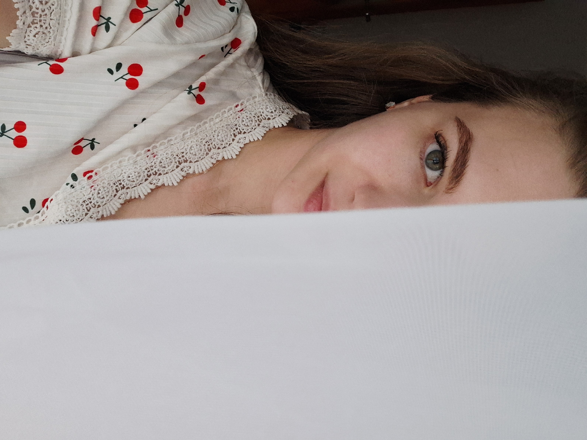 A close-up, slightly blurred, angled eye-level shot captures a portion of a woman lying down. She is partially covered by a white sheet or blanket. Her face is partially visible, showing one eye, an eyebrow, part of her cheek, and her lips. She has fair skin, brown hair, and her eye appears to be green. She is wearing a white top with a cherry print and lace trim around the collar. The background is dark and out of focus. There is no information given about the location in the city of Chaikovsky, Russia.