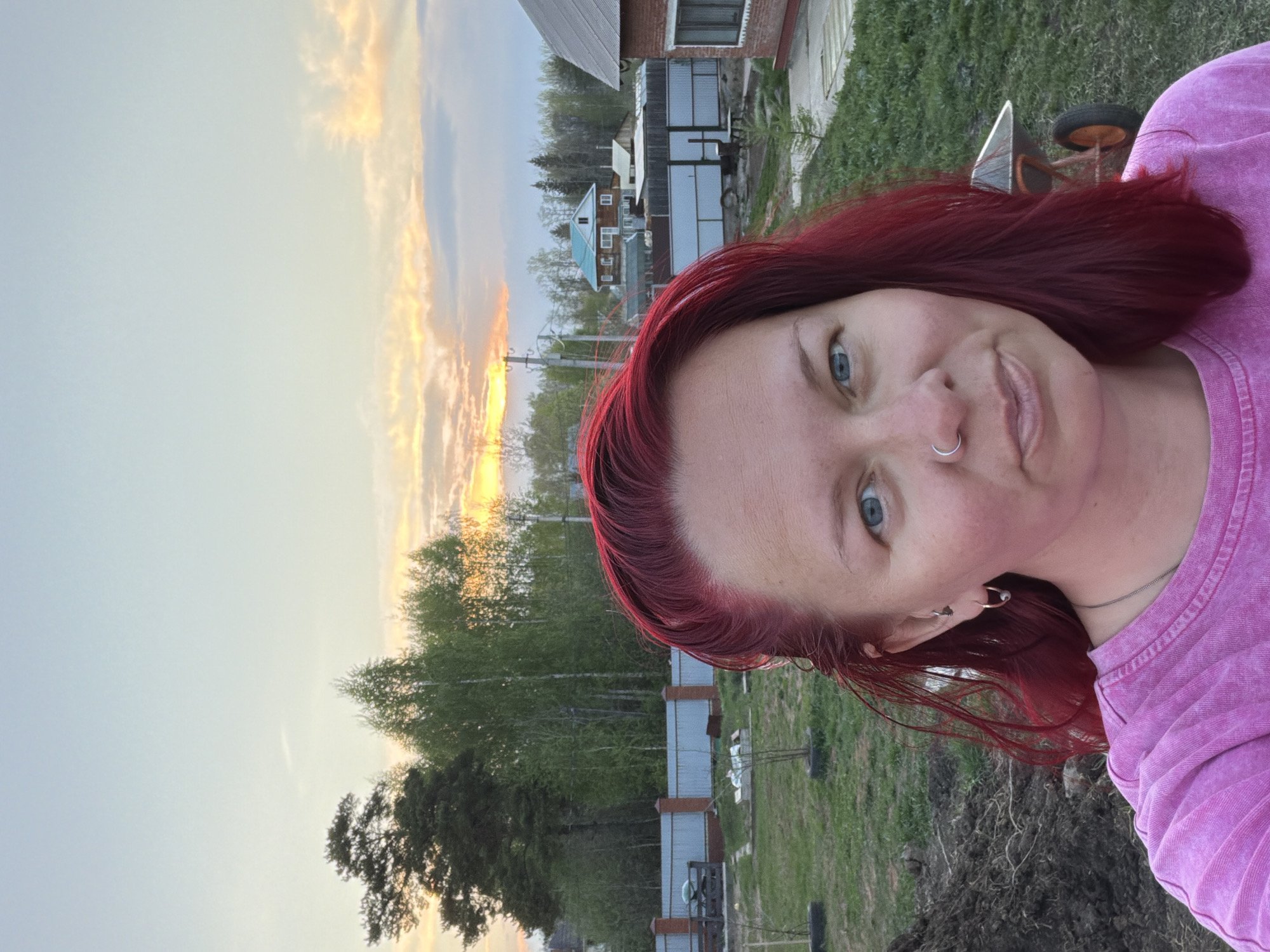 A selfie of a woman with red hair and light blue eyes is oriented vertically. She is wearing a pink shirt. She has a nose ring and multiple earrings. Behind her is a landscape including trees, a setting sun with a multicolored sky, houses, a fence, and a dirt patch in a grassy yard. This photo appears to have been taken in the city of Chaikovsky, Russia.