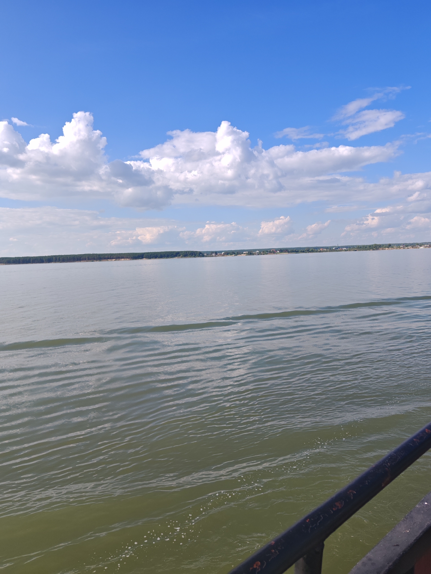 A wide, calm body of water stretches to a distant, tree-lined shore with scattered buildings under a bright blue sky dotted with white, fluffy clouds. In the foreground, a dark metal railing, slightly rusted in places, angles diagonally across the bottom right of the frame, obscuring a portion of the water's surface. The water itself has a greenish hue and exhibits subtle ripples, with a faint wake visible closer to the railing. The horizon is defined by the landmass and the sky, with the water occupying the majority of the image. The scene appears to be a natural landscape, possibly a river or lake, with human settlement visible on the far bank. The location is identified as Nizhnekamenka, Russia.