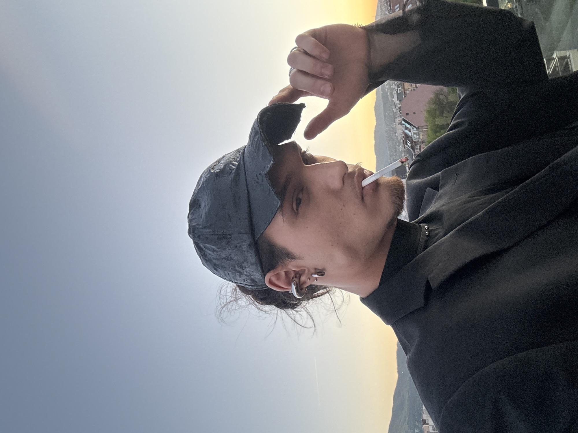 A man is smoking a cigarette. He is wearing a black jacket and cap. He is making a hand gesture over the top of his head, possibly shielding his eyes from the sun. He has facial hair and earrings. The setting appears to be outdoors, possibly a rooftop overlooking a city at sunset. The location is Sofia, Bulgaria. There are buildings and hills in the background.
