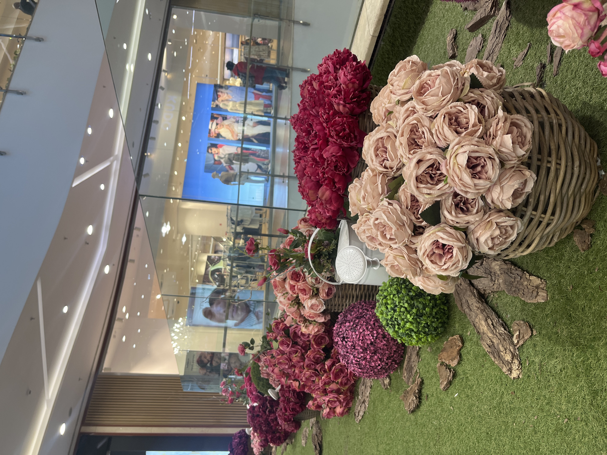 The image depicts a floral display in what appears to be a shopping mall in Sofia, Bulgaria, based on the Cyrillic script visible in the advertising posters.
The display consists of several arrangements of artificial flowers. There are clusters of roses in shades of pink and red, arranged in wicker baskets. A small, white watering can is interspersed among the flowers. These are placed on a base of artificial green grass, and scattered pieces of bark.
The display is positioned adjacent to a large glass wall, which reflects the interior of the mall. Through the glass, people can be seen walking. Advertising posters are affixed to the wall behind the display. The ceiling of the mall is visible at the top of the frame, with rows of lights. The overall setting is a well-lit, modern commercial space.