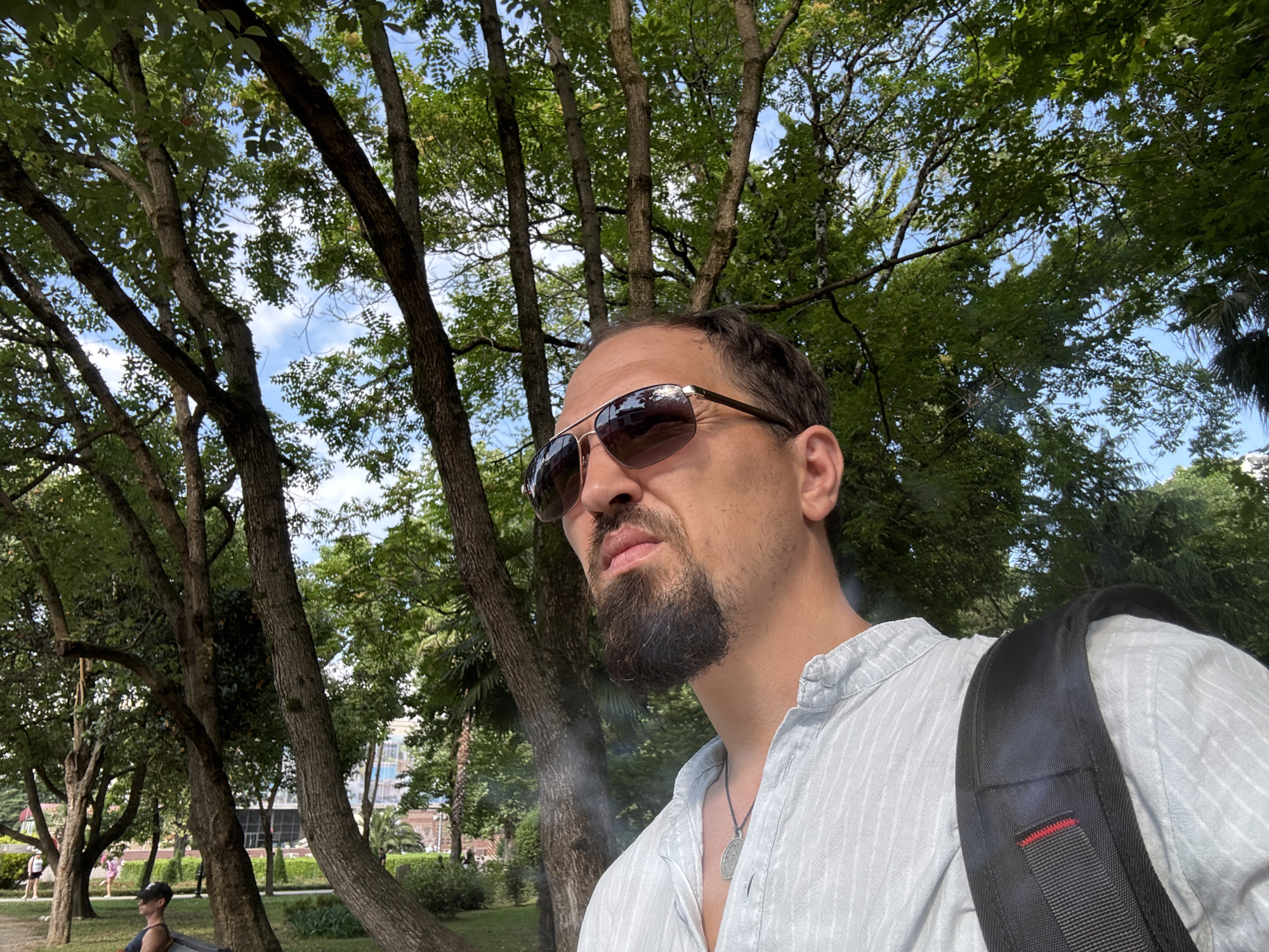 This is a selfie taken outdoors in Sochi, Russia. The subject is a man with a beard and sunglasses, wearing a light blue striped shirt and a backpack. He is looking to his left, with a neutral expression. The background is a park with many trees and foliage. The sky is visible through the leaves, suggesting it is daytime and the weather is clear. In the background, there are hints of buildings and other people enjoying the park. The overall scene conveys a relaxed, casual atmosphere.