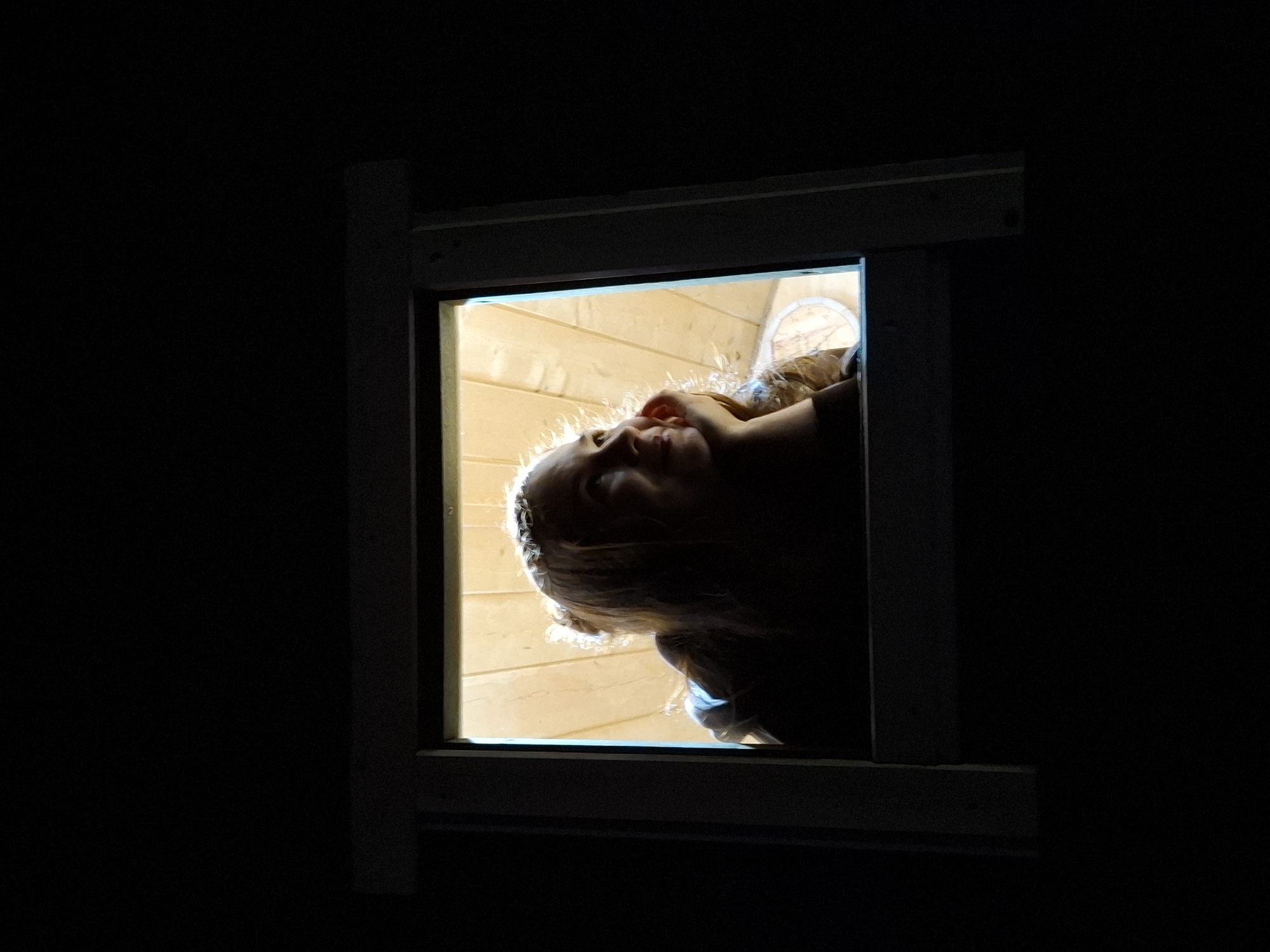 A person with blonde hair looks down through a window. The window frame is white, and the surrounding area is dark. The background seen through the window is light yellow. The person's face is partially obscured by shadows. The location is not discernible from the image.