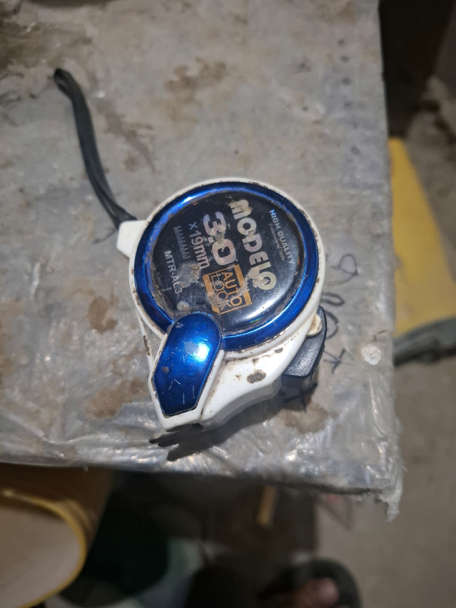 A close-up, top-down shot captures a white and blue tape measure with a black strap resting on a dirty, rough surface. The tape measure, which appears to be from the brand "MODELO," displays "3.0 AUTO LOCK" and "X19mm" on its blue casing. The surface it rests on is textured, possibly concrete or a similar material, with dirt smudges and debris scattered across it. The background is blurred, but hints of a yellow object and plastic sheeting are visible. The scene is set in Kutasirna, Indonesia, as indicated by the provided location.