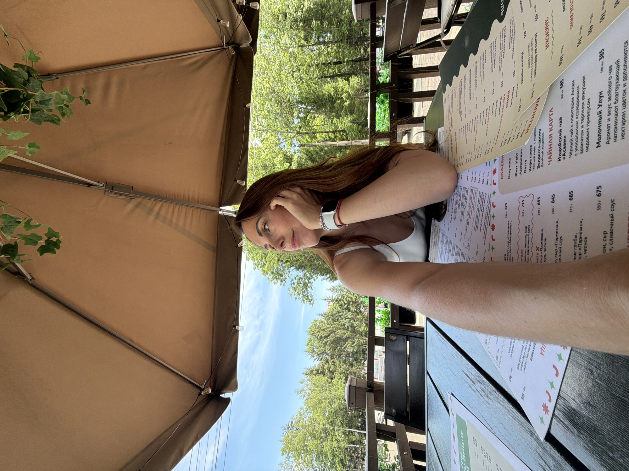 A woman with long brown hair is sitting at a table under a large outdoor umbrella. She is wearing a white tank top and a watch on her left wrist. She is looking to her right, with her hand on her cheek. The table is covered with menus. In the background, there are trees and a fence. The setting appears to be an outdoor cafe or restaurant. The image was taken on a sunny day. The specific location in Birobidzhan, Russia cannot be determined from the image alone.