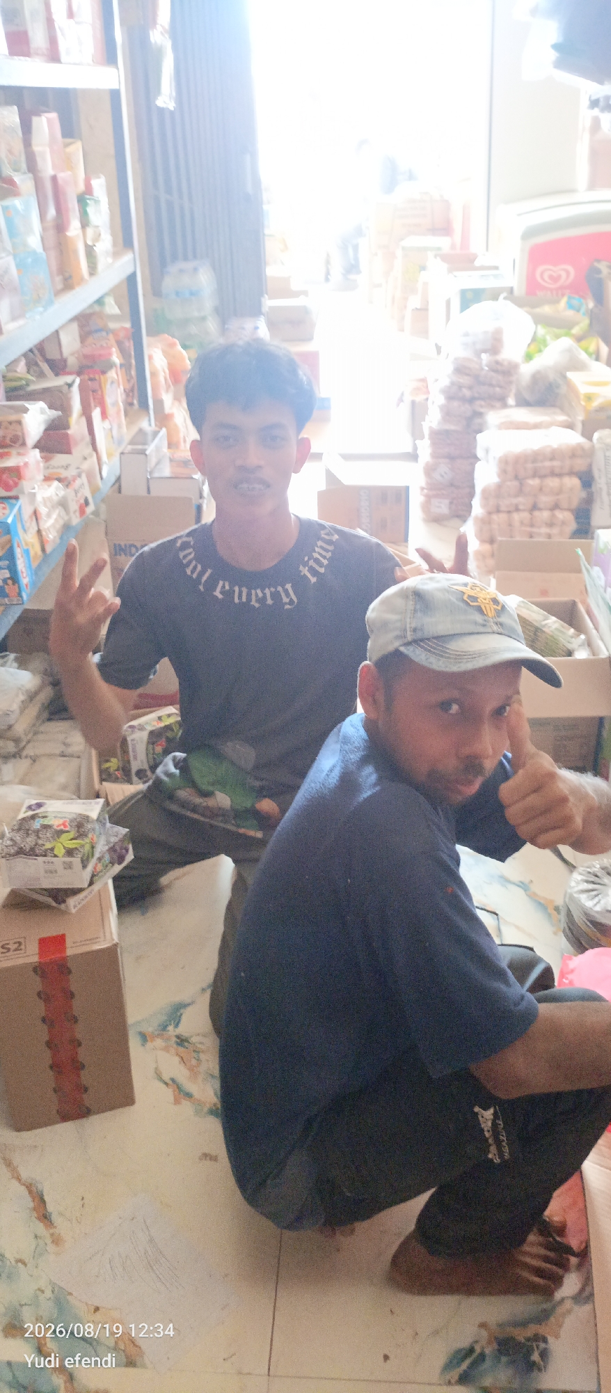 Two men are captured in a photograph, crouching on a tiled floor. The man on the left, wearing a dark grey t-shirt with white text, has his right arm extended upwards, fingers forming a "rock on" gesture. His left hand is also raised, with fingers bent in a similar pose. He has short, dark, curly hair and is looking directly at the camera with a slight smile and braces. The man on the right wears a blue t-shirt and a light blue baseball cap. His right hand is raised, giving a thumbs-up to the camera, and he is looking towards the viewer with a curious expression.
The background suggests an indoor setting, possibly a shop or a storage area, filled with various products stacked on shelves and boxes. Some of these items appear to be packaged food or drinks. The lighting is bright, with a strong light source coming from the right side of the image, creating a glare and partially obscuring details in the background. The floor has a marbled pattern with some colorful splotches. A timestamp and name "2026/08/19 12:34 Yudi efendi" are visible in the bottom left corner of the image.