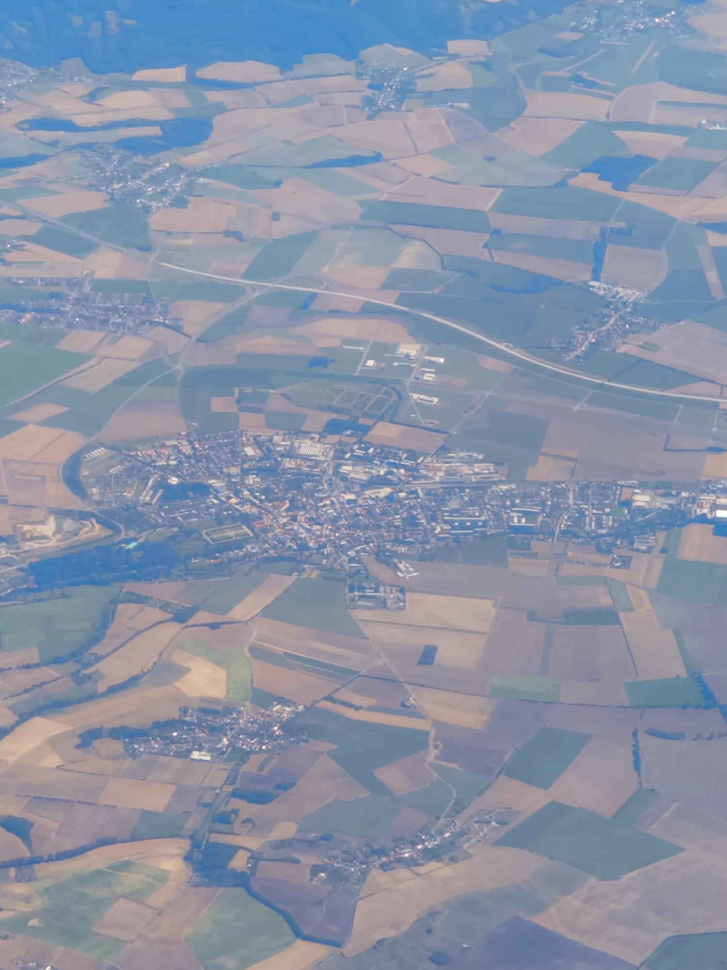 This is an aerial photograph taken from an airplane, looking down upon a landscape. The scene is dominated by a patchwork of agricultural fields, displaying variations in color due to different crops or stages of growth, ranging from shades of brown and tan to green and purple. A winding river or waterway is visible, cutting through the terrain, with a settlement nestled along its banks. The settlement appears to be a town or village, characterized by a dense cluster of buildings with red-tiled roofs and white walls. A road network is integrated into the landscape, connecting the settlement to surrounding areas and traversing the fields. The overall environment suggests a rural or semi-rural setting, likely in a European country given the architectural style. The location is identified as Radslavice, Czechia. There are no individuals or specific actions immediately discernible from this high-altitude perspective.