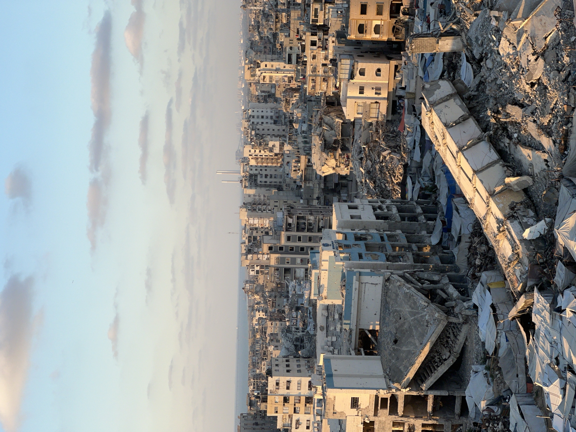 The image depicts a wide aerial view of a heavily damaged urban landscape in Gaza City, Palestinian Territories, under a partly cloudy sky. The scene is characterized by widespread destruction of multi-story buildings, with many structures reduced to rubble or showing significant structural damage. Debris from collapsed buildings fills the spaces between the remaining structures, creating a chaotic and devastated environment.
In the foreground, a large concrete beam lies diagonally across a pile of rubble, indicating recent structural failure. Surrounding this are numerous temporary shelters, appearing as white and blue tarps or tents, suggesting displacement of residents and makeshift living arrangements. The damaged buildings exhibit exposed concrete, twisted metal rebar, and gaping holes where windows and walls once stood. Some buildings still stand partially intact, but with clear signs of impact and partial collapse.
Further in the background, the cityscape extends towards the sea, with more damaged buildings and in the distance, industrial structures with smokestacks are visible. The overall impression is one of immense destruction and the aftermath of conflict. The lighting suggests it is either early morning or late afternoon, with long shadows cast across the scene. No people are clearly visible in the image, but the presence of temporary shelters implies human activity and habitation amidst the devastation.