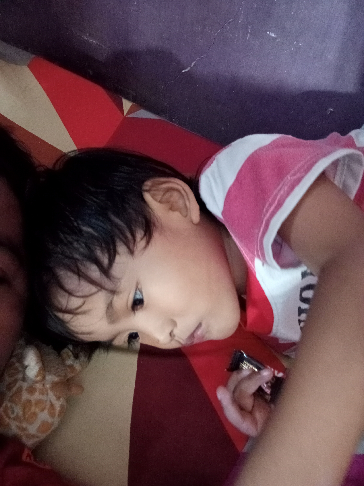The image captures a close-up, overhead view of a young child lying down. The child has dark hair and is wearing a pink and white striped shirt. Their face is turned to the side, and they appear to be looking at something off-camera. A hand is visible, holding a small, dark-colored object, possibly a candy bar. The child is lying on a surface with a red and beige striped pattern. In the background, a dark purple wall with some cracks is visible. The image seems to be taken indoors. Based on the provided context, this scene is located in Bloro, Indonesia.