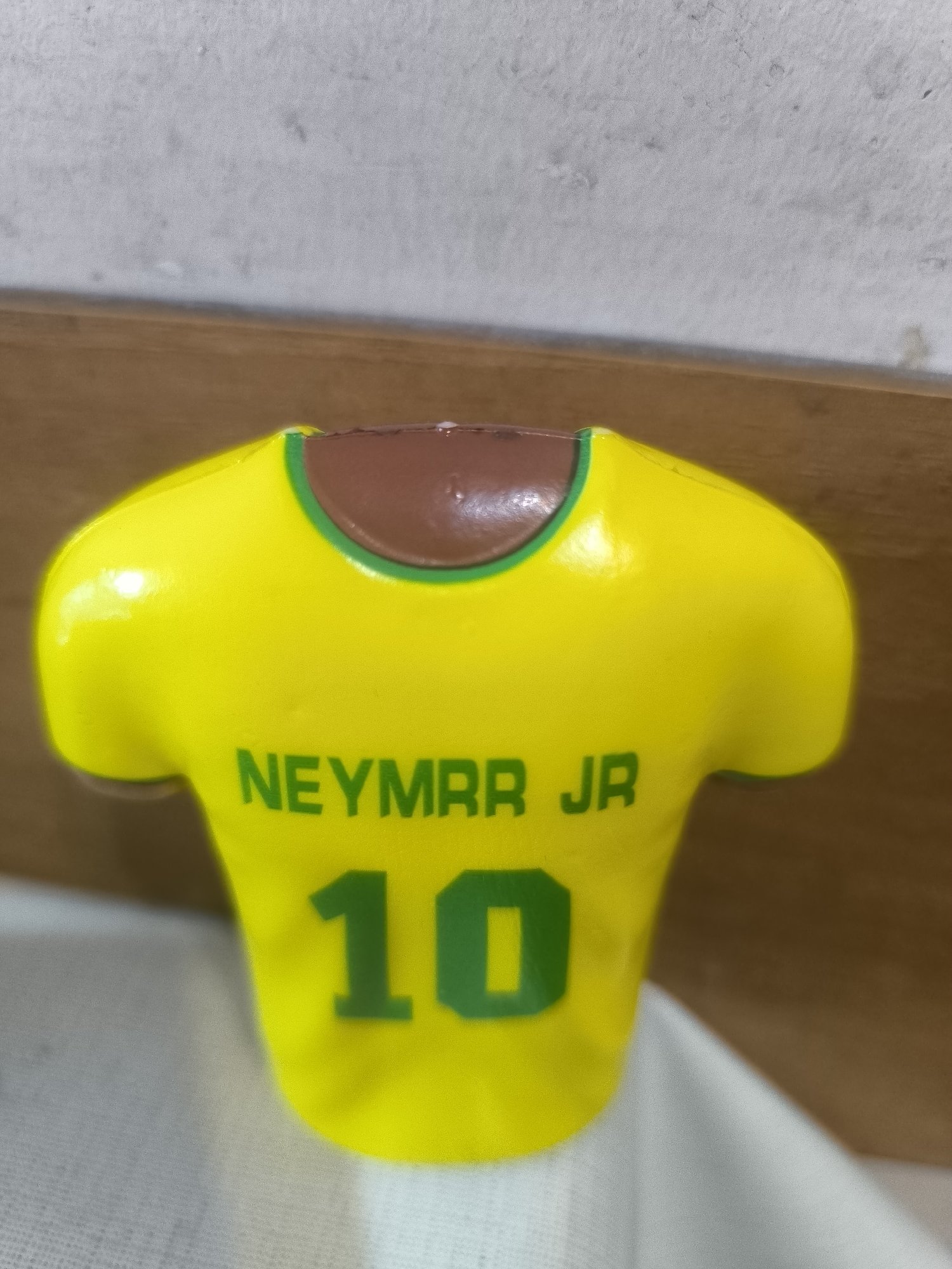 The image displays a close-up of a small, decorative item shaped like a bright yellow soccer jersey. The jersey features a green trim around the neck, which transitions into a brown, molded section suggesting a neck or body beneath the shirt. Prominently printed on the back of the jersey in green capital letters is "NEYMARR JR" above the large number "10".
The item appears to be made of a glossy plastic or similar synthetic material, reflecting some light. It is positioned against a simple background consisting of a light grey, textured wall at the top, and a horizontal wooden surface (possibly a shelf or table) running across the middle. A white, textured fabric, likely a cloth or tablecloth, is visible in the foreground, partially obscuring the base of the jersey item. No specific activity or event is taking place; it is a still life photograph of the object indoors in Yerevan, Armenia. The lighting suggests an indoor setting, without clear indicators of time of day.