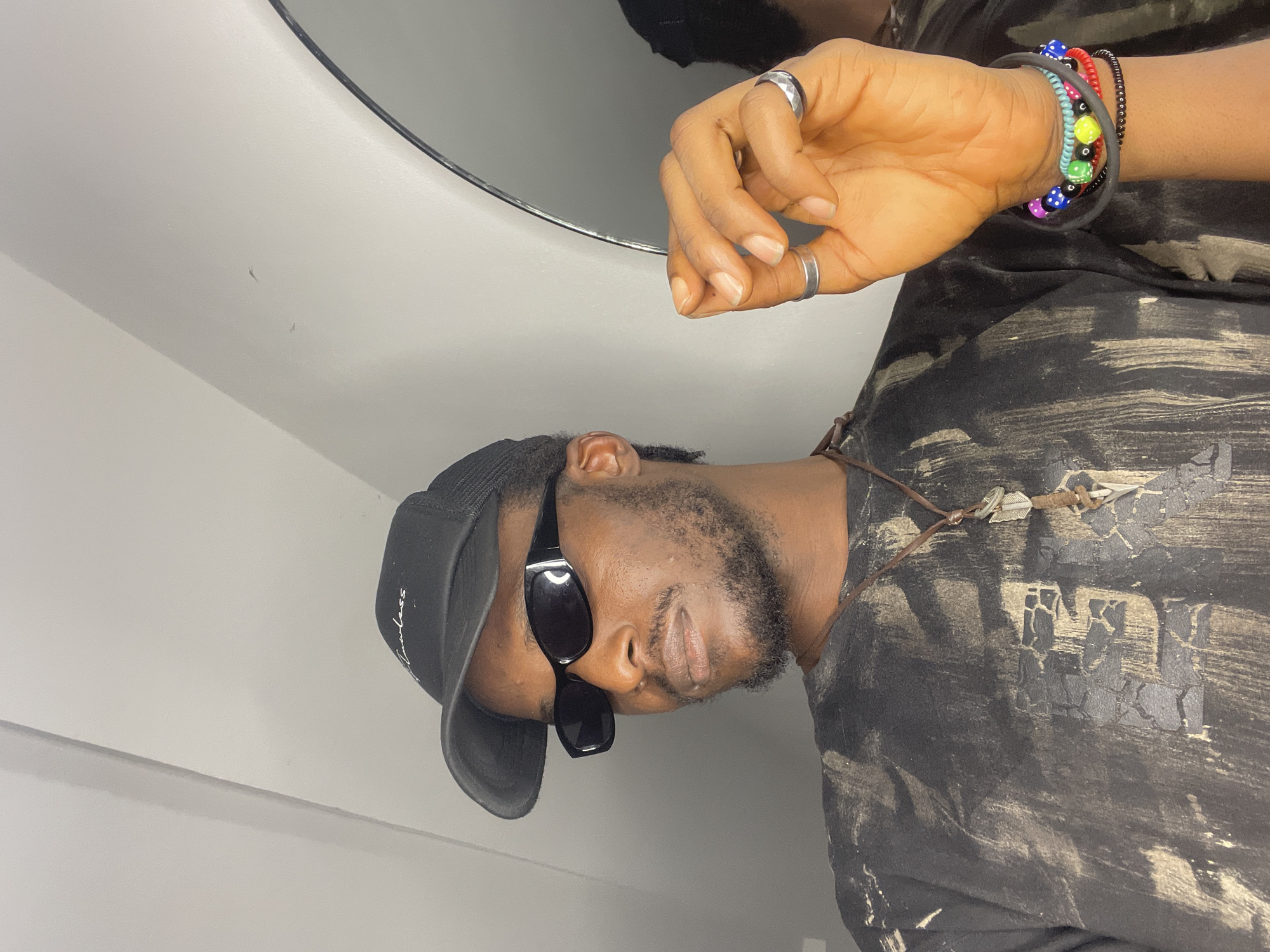 The image shows a close-up of a young Black male wearing a black baseball cap with "Flawless" written on it, black sunglasses, a black t-shirt with a tie-dye or distressed pattern, and a necklace with a pendant. He is also wearing rings on his fingers and multiple beaded bracelets on his wrist. His right hand is raised, with his index finger and thumb touching. The background appears to be an indoor setting with a light-colored wall and a portion of a round mirror reflecting the person. There is no visible indication of the specific location within Abuja, Nigeria.