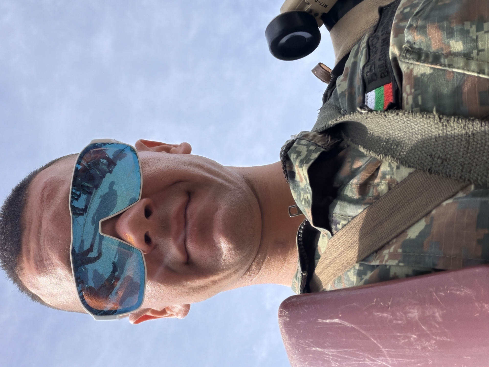 The image is a close-up, low-angle shot of a man wearing camouflage clothing and reflective sunglasses. The man's face is partially obscured by the sunglasses, which reflect a group of people standing on a paved surface. He is wearing a tactical vest or harness, and a patch on his uniform displays the Bulgarian flag and the words "BULGARIAN ARMY." A pink object, possibly a piece of equipment, is visible on his left. A black scope or device is attached to his right. The background is a pale blue sky with some clouds. The scene appears to be outdoors, under daylight conditions. The specific location in Veliko Tarnovo, Bulgaria is not discernible from the image.