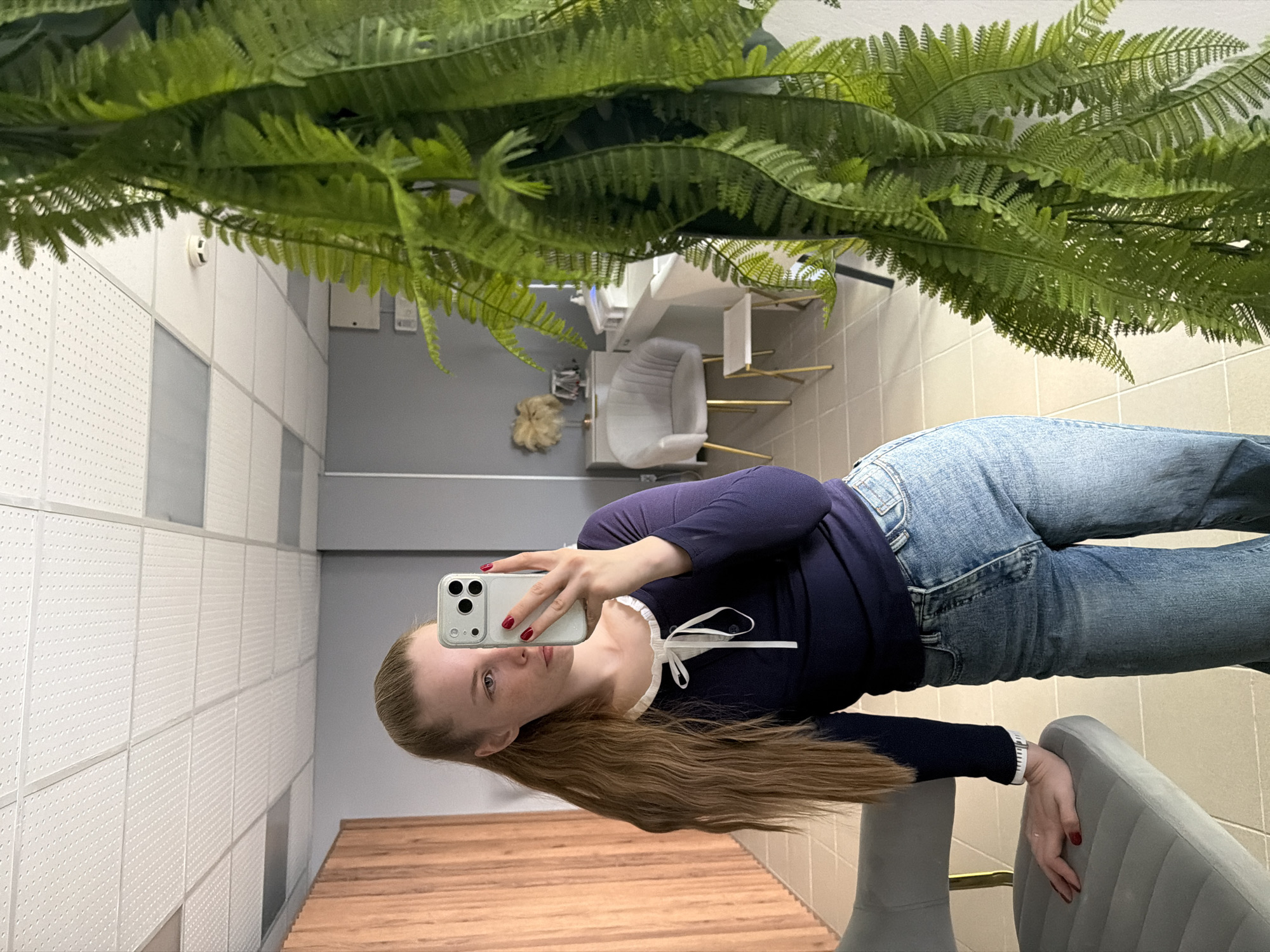 A person is taking a mirror selfie in what appears to be an office setting. The person is wearing jeans and a navy blue, long-sleeved shirt with a white ribbon detail. They are holding a smartphone in their left hand. A fern plant is visible at the top of the frame, hanging from the ceiling. The office includes a desk with chairs, and the walls are tiled. It is not possible to definitively determine the location is in the city of Berezniki, Russia from the visual information provided.