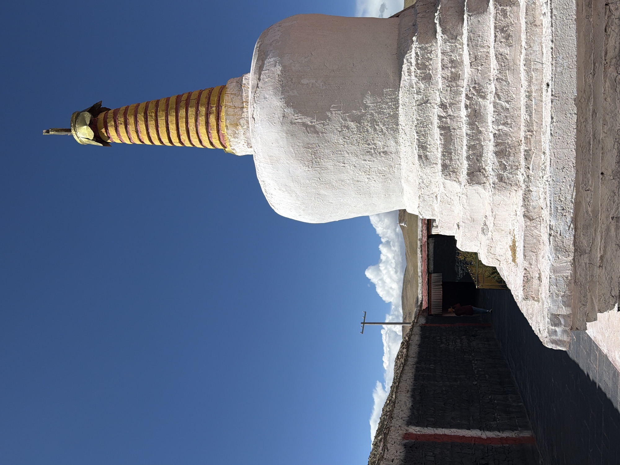 A photograph taken in Labrang township, China, captures a large, white Buddhist stupa (chorten) under a clear, bright blue sky. The stupa dominates the left side of the frame, showcasing its distinctive architecture. Its main body is a bulbous, white structure with a rough, textured surface, indicating it's likely made of whitewashed stone or plaster. Above the dome, a tall, cylindrical finial rises, painted in horizontal bands of gold and dark red. The finial is topped with a small, intricate golden ornament.
To the right of the stupa, the image reveals part of a building with stepped, white walls, suggesting a traditional architectural style. Below these white walls, a dark-colored stone wall with a distinctive red stripe runs horizontally across the midground. An open doorway in this wall shows a person partially visible inside, wearing a dark top and what appears to be a red or maroon jacket. They seem to be resting or seated.
In the background, a wooden utility pole with crossbeams stands against the bright sky, with scattered white clouds visible above distant, rolling hills or mountains on the horizon. The lighting suggests it is bright daylight, likely midday or early afternoon, with clear weather and distinct shadows. No discernible text is visible in the image. The overall scene conveys a sense of peacefulness and cultural significance typical of Tibetan Buddhist sites.