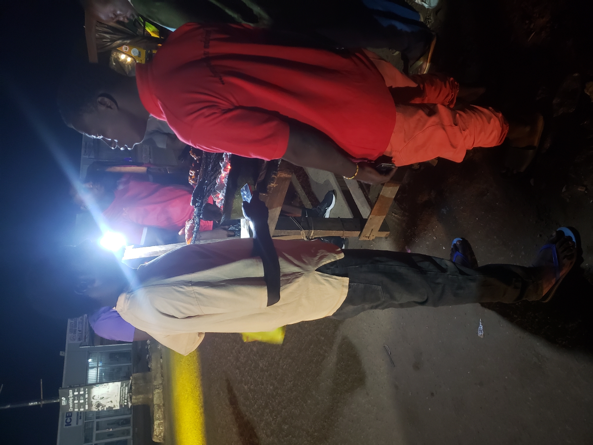The image, taken at night in what appears to be a street or market setting, shows several individuals gathered around a food stall or grill. The scene is illuminated by harsh artificial light sources, creating strong highlights and shadows, with significant lens flare in the lower-left quadrant. The photograph is oriented vertically, showing people standing upright from a sideways perspective.
In the foreground, a person in a white or light-colored short-sleeved shirt and dark pants stands with their back mostly to the viewer, holding a smartphone. This individual is wearing blue flip-flops. To their right, closer to the center, another person is prominent, wearing a red long-sleeved shirt and orange pants. This individual has a gold-colored bracelet on their right wrist and appears to be interacting with the food stall. Behind them, a dark, possibly grilled item on skewers is visible, suggesting food preparation or sale. Another person in a pink or maroon shirt is partially visible behind the person in red.
The ground is dark asphalt or concrete, with a small knife visible on it in one of the lower crops. In the background, further to the left, a building with various signs is visible. One sign clearly displays "ICE Co-working Space." The overall atmosphere suggests a bustling night street scene, possibly involving street food vendors and customers. Multiple individuals are using smartphones, either for communication, light, or other purposes.