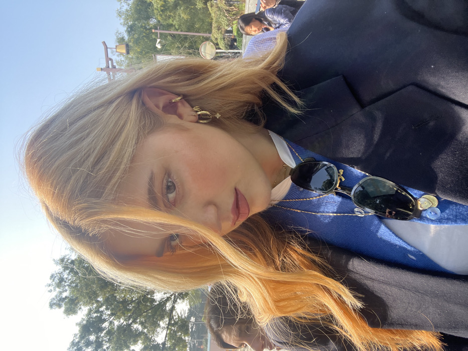 A young adult female with long, wavy golden-blonde hair and light-colored eyes is captured in a medium close-up, looking directly towards the camera. Her visible right ear features multiple gold-colored hoop earrings, one with a star charm, and a cuff earring. She is dressed in a dark navy blazer over a blue V-neck cardigan and a white collared shirt. Dark-tinted sunglasses are suspended from a gold-colored chain around her neck, resting on her chest, alongside additional gold necklaces with various charms. The outdoor background, under bright daylight, includes dense green foliage and a traditional Chinese-style wooden structure with a bell, consistent with architecture found in Lintong District, China. Partially visible individuals are present in the blurred background.