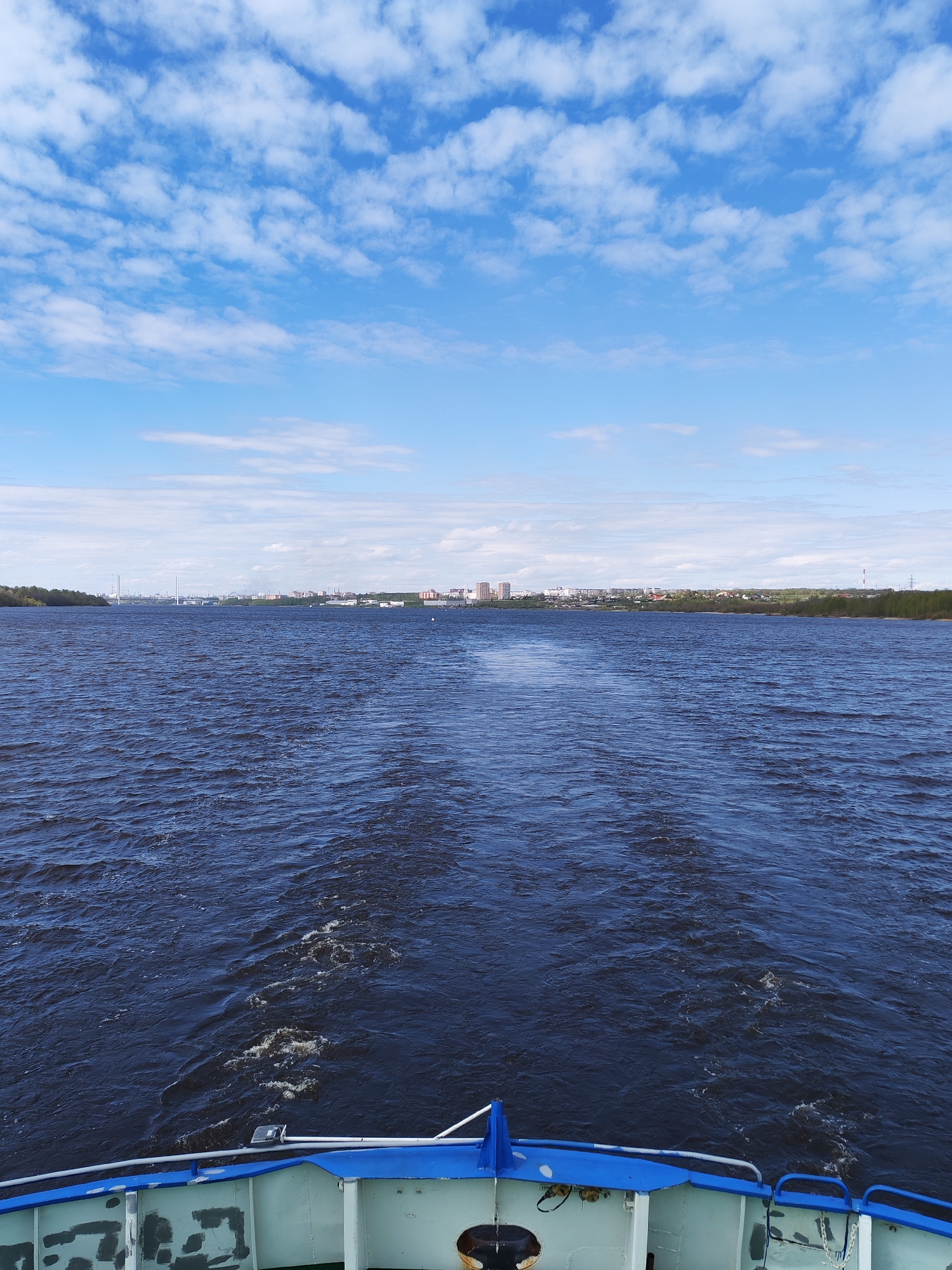 The image captures a view from the stern of a vessel moving across a body of water. The water is dark blue and disturbed by the wake of the vessel. The sky above is a bright blue with scattered white clouds. In the distance, a cityscape stretches along the horizon, with buildings and infrastructure partially visible. The stern of the vessel is visible in the foreground, showing a blue and white painted structure with some fixtures. The location is Unknown, Unknown.