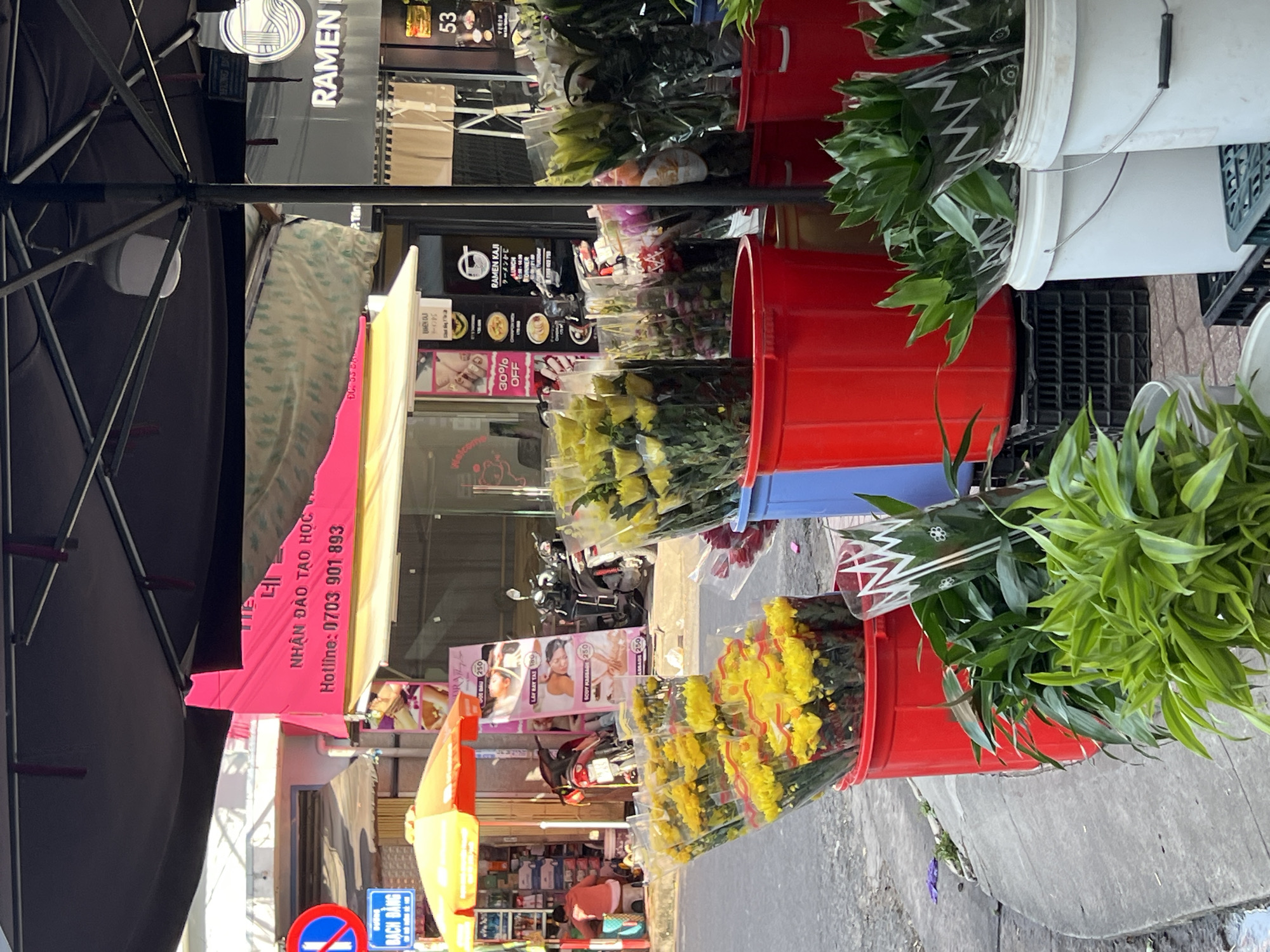 This image depicts a street scene in Nha Trang Ward, Vietnam, focusing on a flower stall. The foreground is dominated by several large red buckets filled with bouquets of yellow chrysanthemums, some of which are wrapped in clear plastic. To the right, white buckets contain bunches of green leafy plants. Behind the flowers, a pink banner is visible with Vietnamese text, including a hotline number. Further in the background, shops are lining the street, one of which displays a "RAMEN KAJI" sign. A yellow umbrella shades a portion of the street, and several motorbikes are parked nearby. The environment appears to be a bustling urban setting, with a sidewalk and street visible.