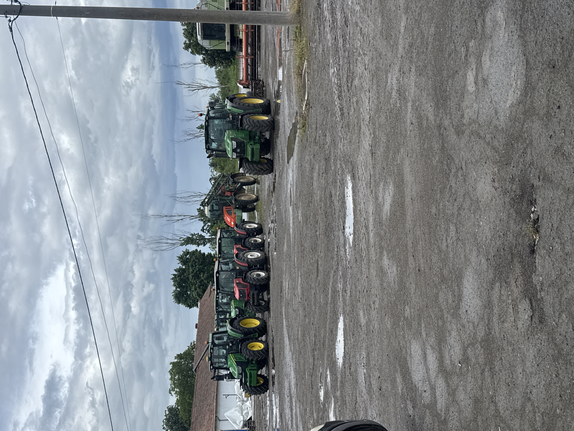 The image depicts a row of tractors parked in what appears to be a lot with a building visible on the left edge, under a cloudy sky. The tractors are a mix of green and red colors, with yellow wheels on some of the green tractors. The ground is wet, possibly from recent rain, with puddles visible. The image suggests a rural setting, potentially in or near Sarnevo, Bulgaria. There are utility poles and wires visible. It's impossible to discern any interactions or specific actions without more information.