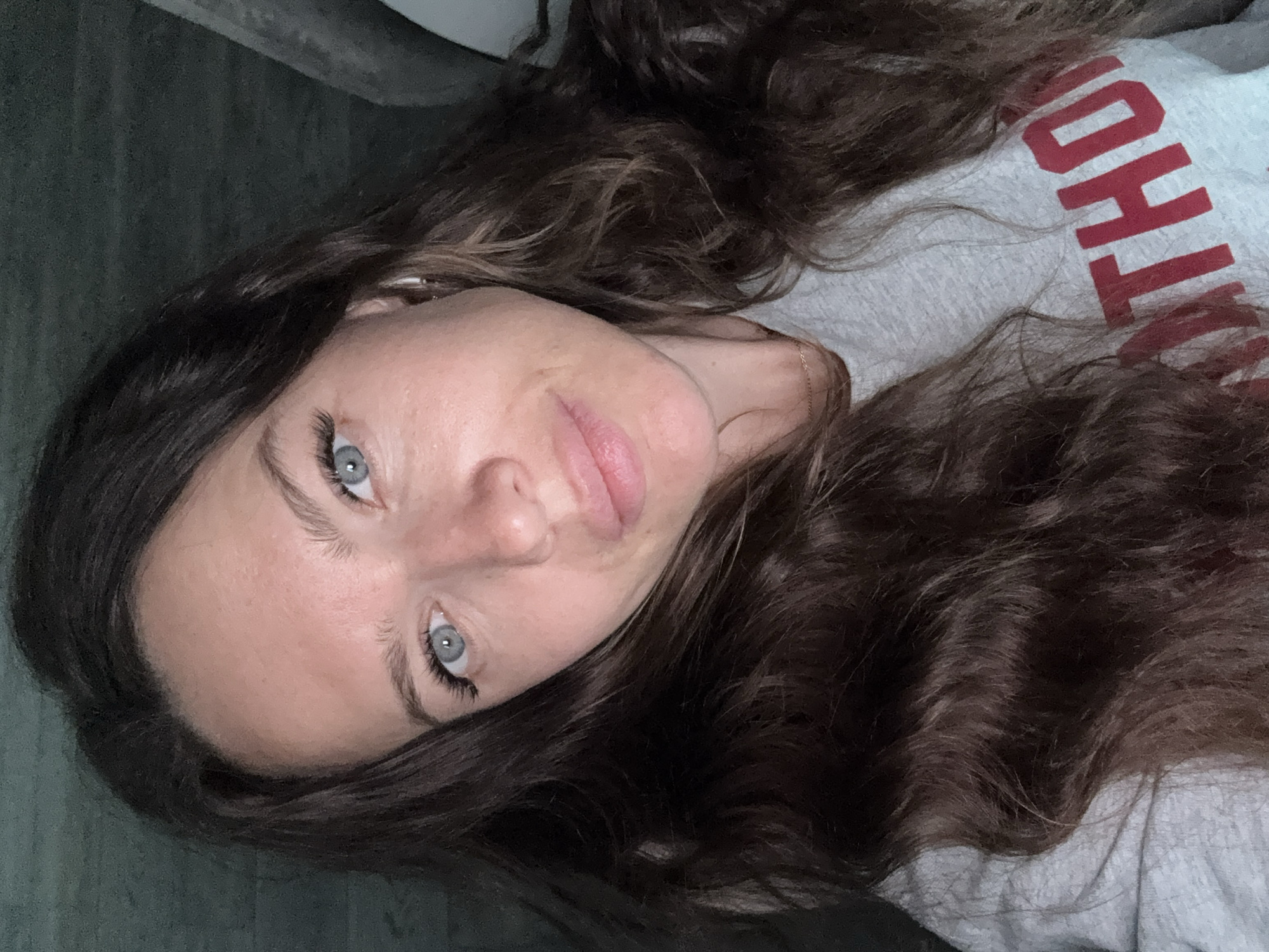 A young woman with long, dark curly hair and light blue eyes is shown in a close-up selfie. She is wearing a light gray sweatshirt with red lettering, partially visible. The background is dark and indistinct. There is no evidence of the location being Dolgoprudny, Russia.