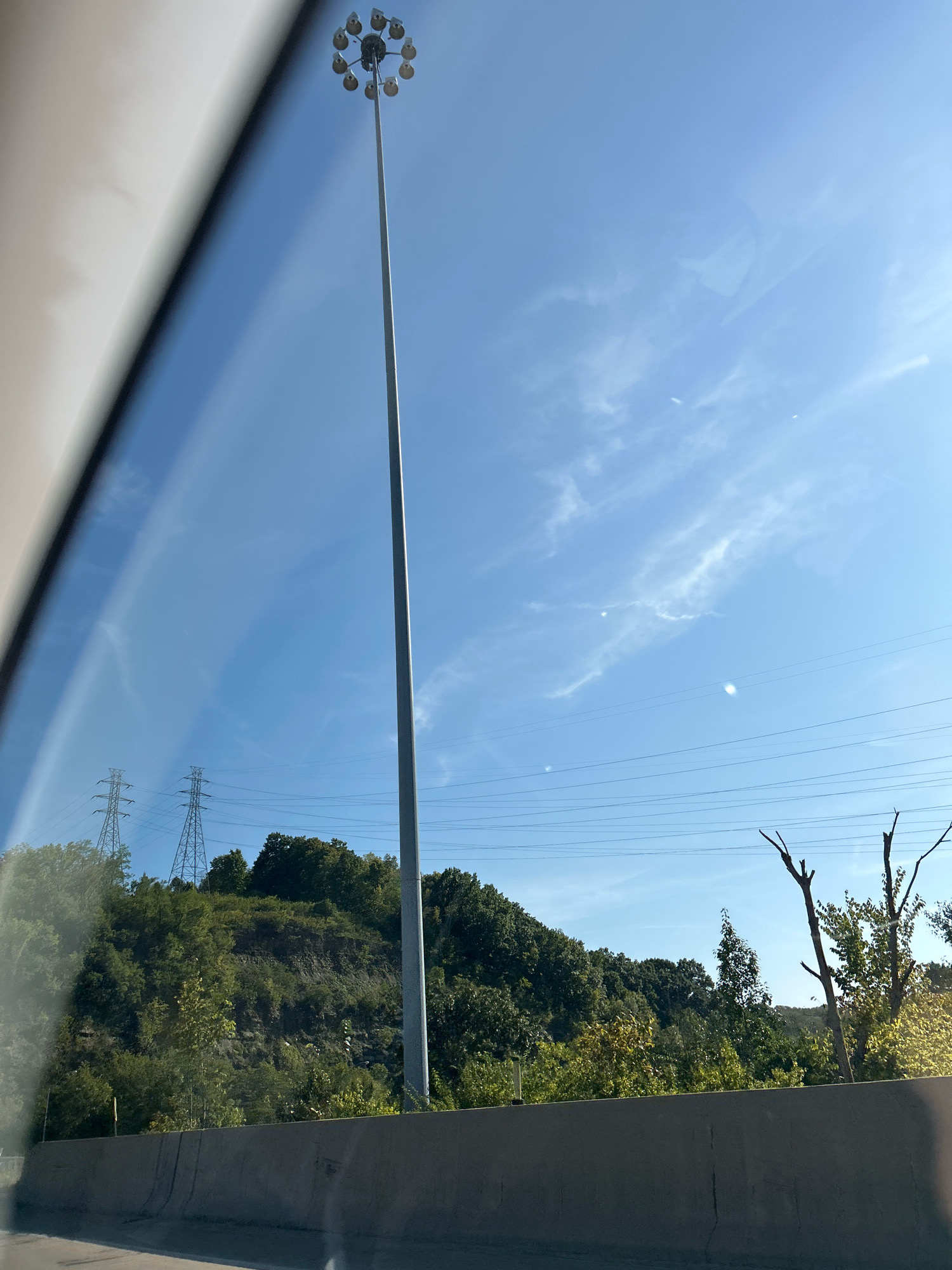 A tall, silver, multi-headed light pole stands prominently against a bright blue sky with wispy white clouds. Power lines crisscross the sky, supported by large, latticed towers on a densely wooded hill in the background. The hill is covered in a variety of green trees and vegetation, with a rocky embankment visible in some areas. In the foreground, a concrete barrier runs horizontally across the bottom of the image, suggesting the photo was taken from a road or highway. The lighting indicates a sunny day. The scene is likely an outdoor, possibly urban or suburban, setting.