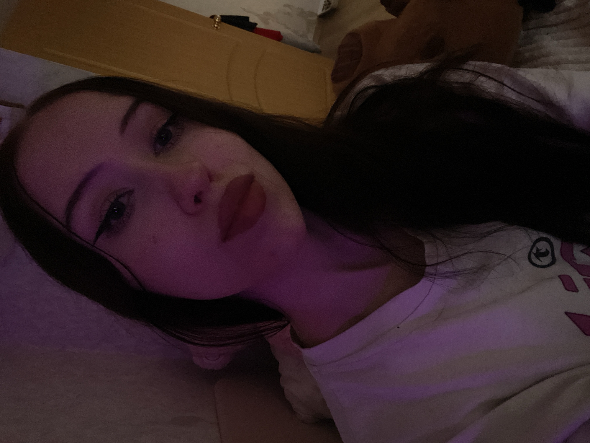A young woman with dark hair and makeup is pictured indoors. She is wearing a white t-shirt. The room is dimly lit with purple and yellow lighting. A wooden door is visible in the background. The scene is likely a bedroom in Votkinsk, Russia.