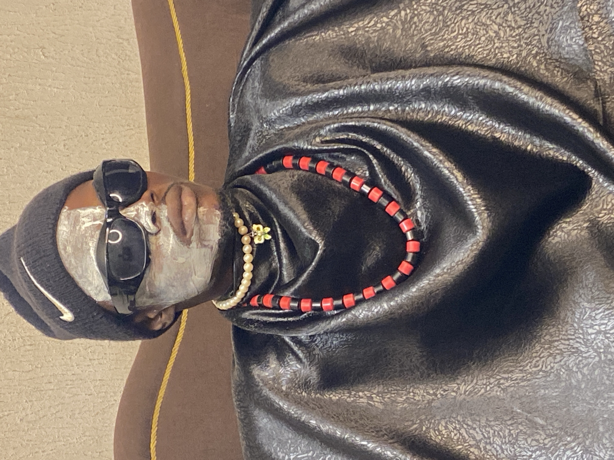 A person is seated on a brown couch in what appears to be an indoor setting, possibly in Abakaliki, Nigeria, given the request. The individual is wearing a black Nike beanie, dark sunglasses, and a shiny black garment that resembles a cape or poncho. Their face is partially covered with a white substance, likely a face mask. They are accessorized with two necklaces: one appears to be made of pearls with a small green charm, and the other is a strand of red and black beads. The background consists of a textured wall. The lighting suggests it might be a daytime shot.