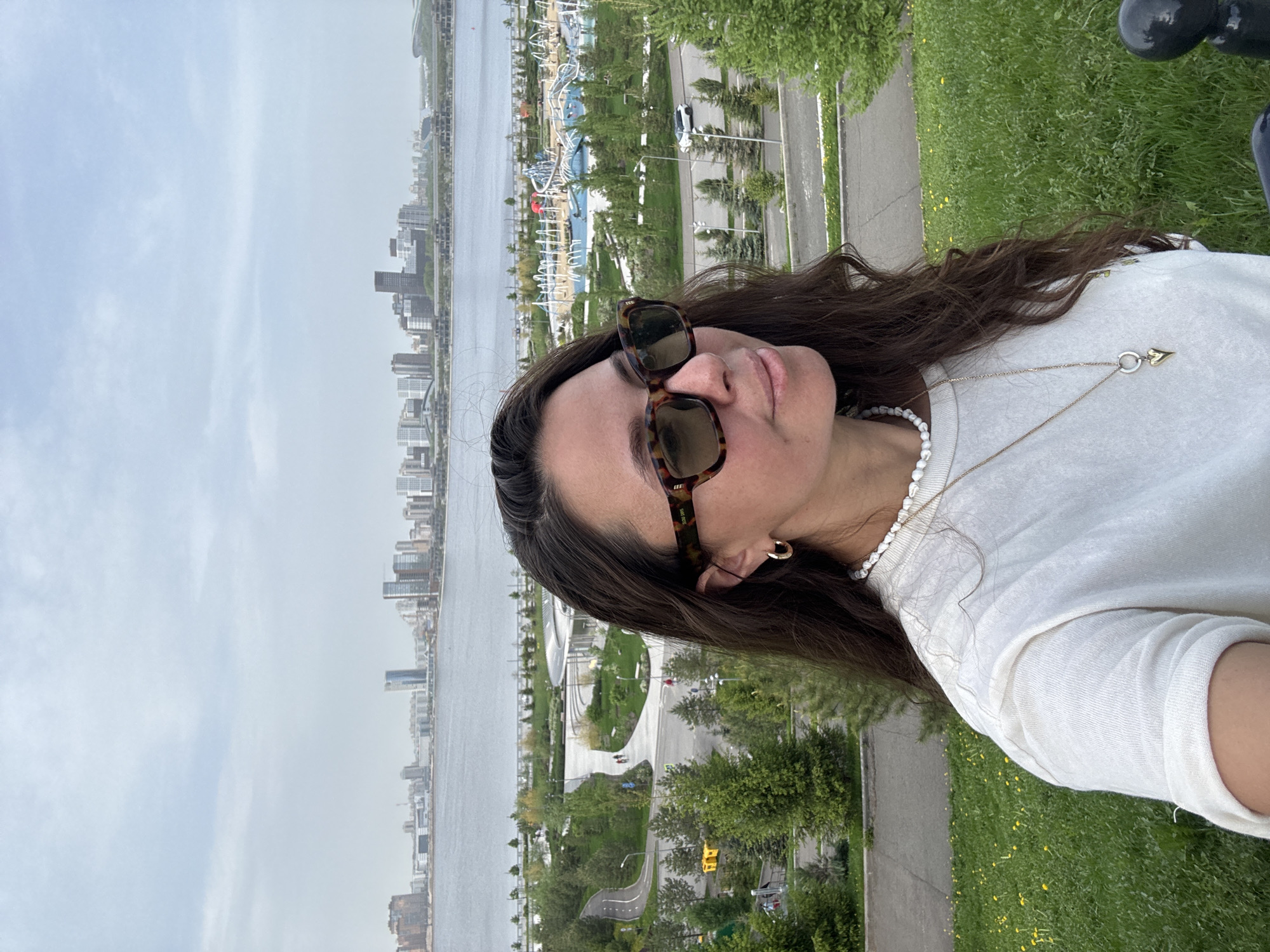 The image depicts a woman wearing sunglasses, a white shirt, and two necklaces with a cityscape in the background. The cityscape includes a body of water, buildings, a park with paths and playground equipment. Based on the cityscape, the location is likely near the Kazan Kremlin embankment, along the Kazanka River in Kazan, Russia. The woman appears to be taking a selfie or is being photographed in an elevated location overlooking the city and river.