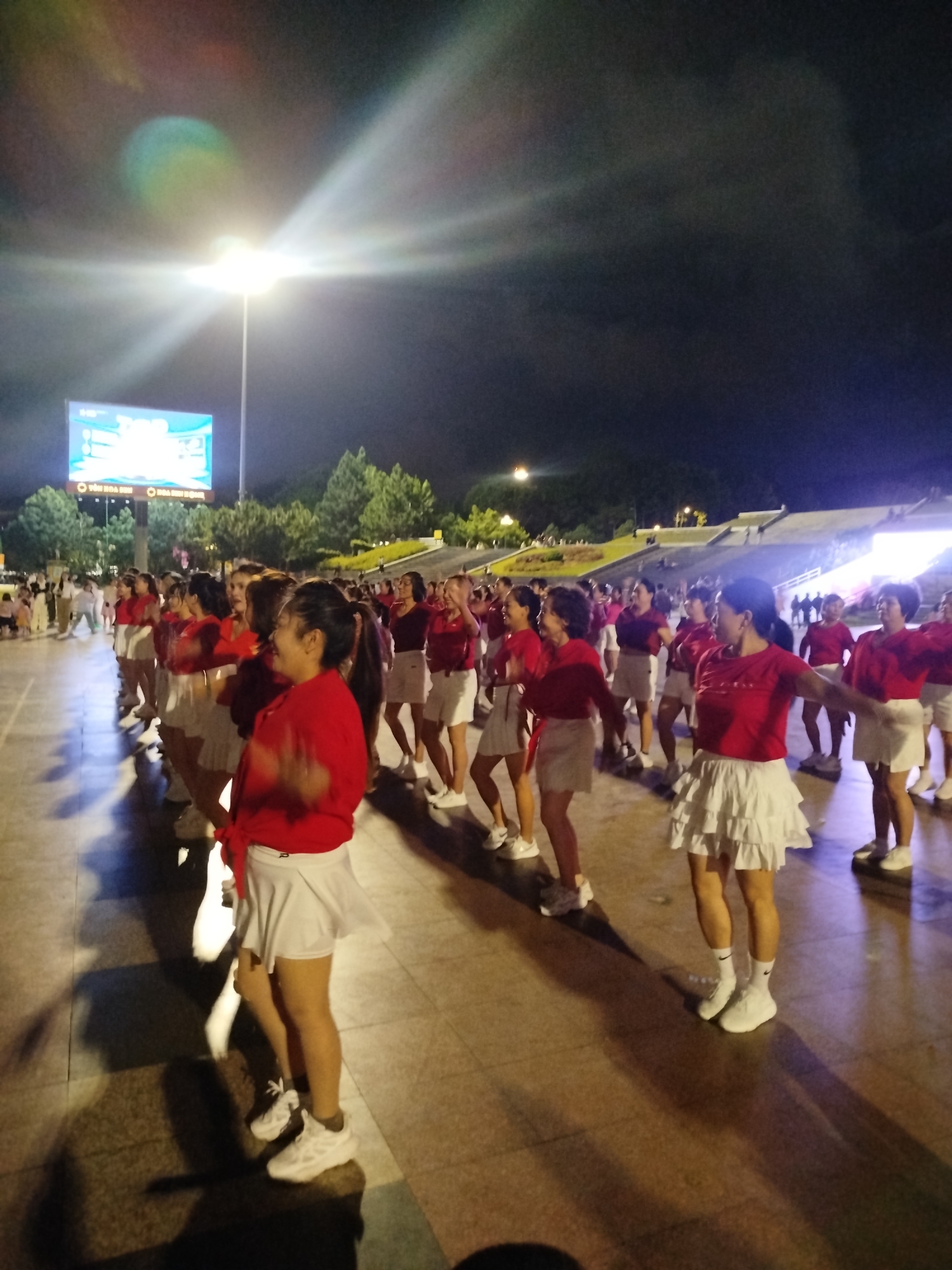 A group of people, primarily women, are engaged in what appears to be a synchronized dance performance at night. They are dressed in matching red tops and white skirts, with white athletic shoes. The scene is outdoors, in a paved area with trees and a large tiered structure in the background, possibly an amphitheater or viewing area. A large LED screen is visible in the distance, displaying text. The lighting suggests it is nighttime, with bright artificial lights illuminating the area. The mood appears to be energetic and festive, with some individuals smiling. The location is Da Lat, Vietnam. The text on the LED screen is partially visible and appears to be in Vietnamese.