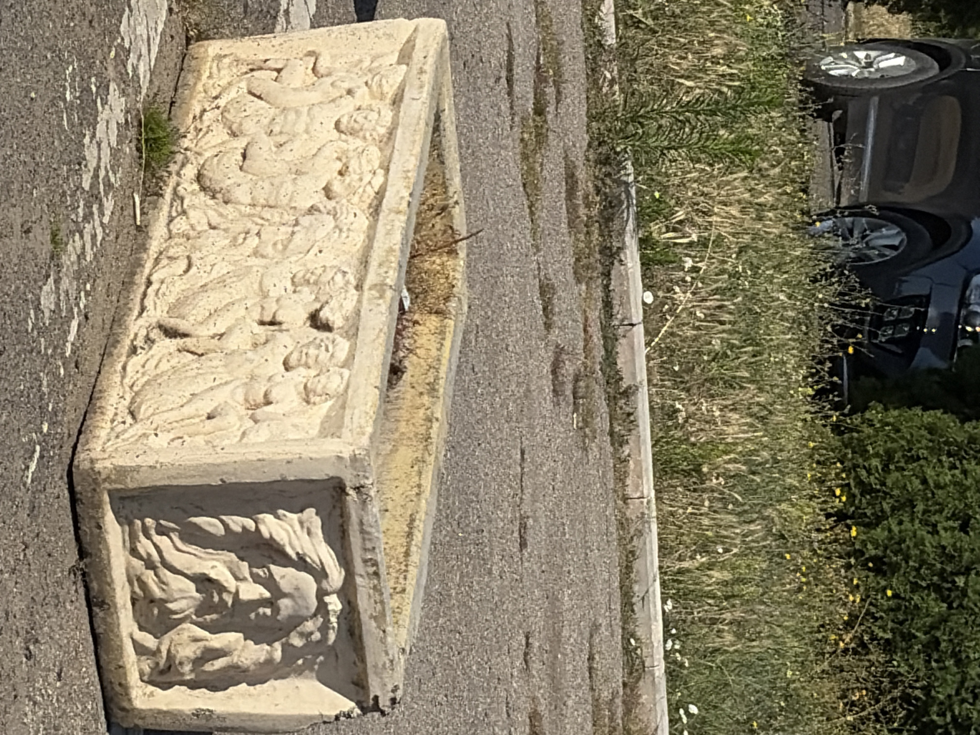 The image shows a weathered rectangular stone planter with relief carvings on its sides. One side features a procession of figures, likely mythological or allegorical, depicted in classical style with draped clothing. The other visible side displays a detailed relief of a bearded male face with a stern expression, framed by flowing hair or drapery. The planter is positioned on an asphalt surface with scattered debris and small patches of grass. In the background, a dark-colored car is partially visible, parked beside a border of overgrown grass and vegetation with small yellow and white wildflowers. The overall scene suggests an outdoor urban or park setting in Criuleni, Moldova, possibly a public space where the planter serves as a decorative or functional element.