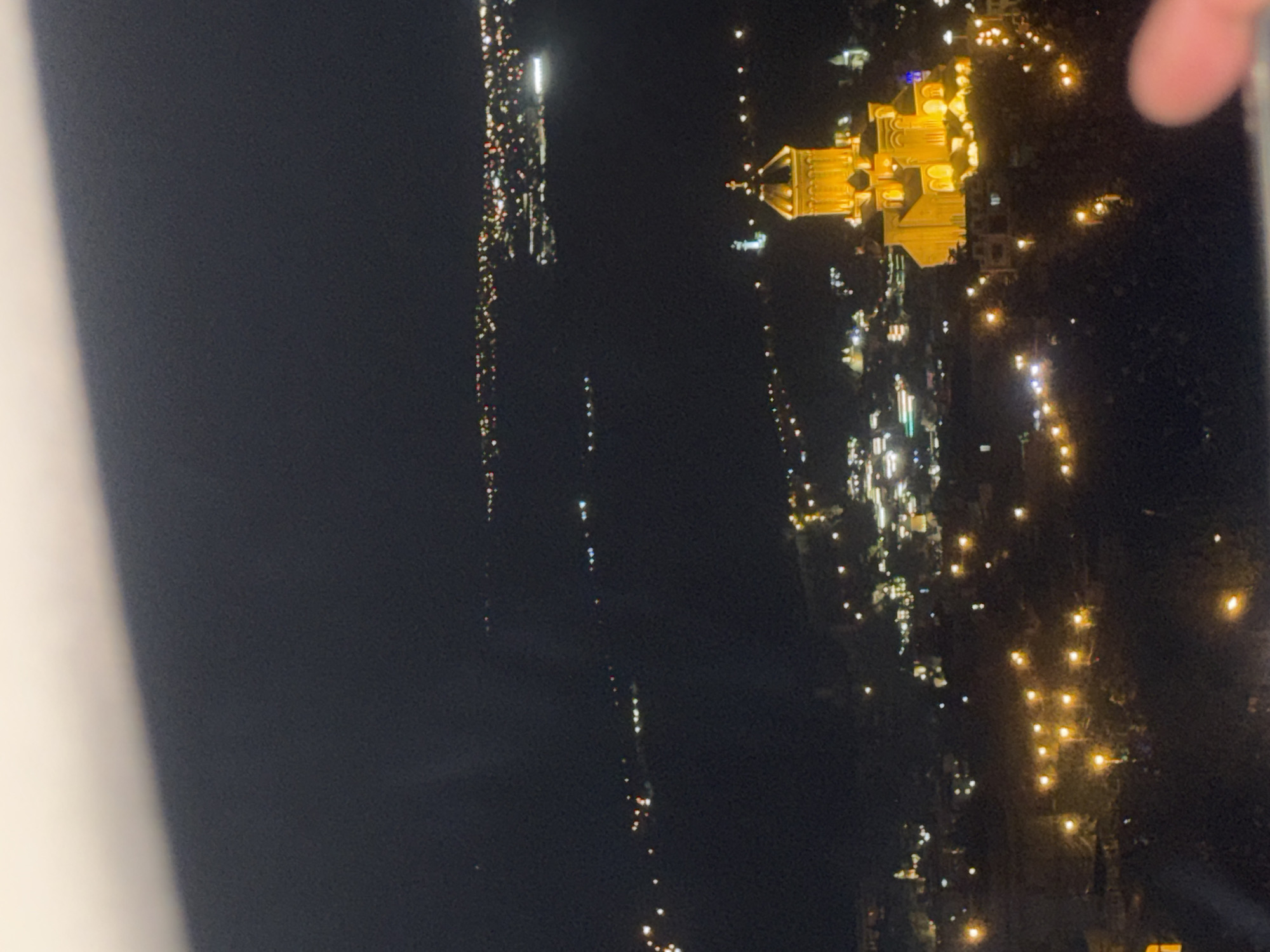 The image shows a nighttime view from an elevated position, likely a high-rise building or a mountain overlooking the city of Tbilisi, Georgia. The dominant feature is the illuminated Sameba Cathedral (Holy Trinity Cathedral of Tbilisi), visible in the upper-right quadrant, glowing with a bright yellow-orange light. A line of bright lights stretches into the distance, representing a street or avenue extending away from the cathedral. Additional clusters of lights suggest residential or commercial areas. The left side of the image is predominantly dark, possibly showing less populated areas. A white vertical element is visible in the left-hand side. A fingertip can be seen in the upper right corner. The overall scene depicts the urban landscape of Tbilisi at night, highlighted by the architectural grandeur of the Sameba Cathedral.