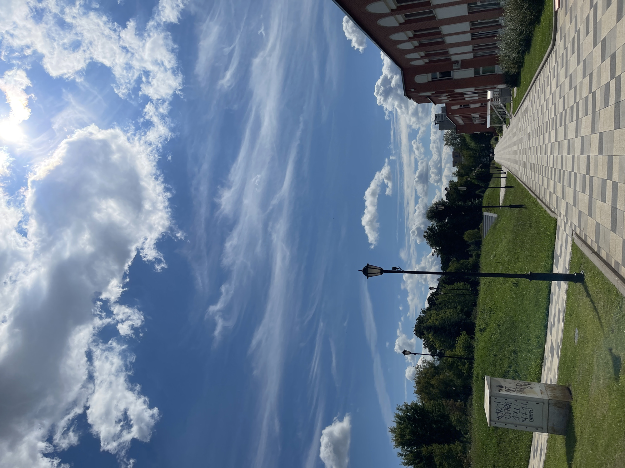 The image depicts a clear, sunny day with scattered white clouds against a bright blue sky. A paved walkway, laid with alternating light and dark square tiles, stretches into the distance, bordered by a green grassy slope on the left and a red brick building with white arched windows on the right. Several lampposts are visible along the walkway. Trees and foliage line the background, with more buildings faintly visible further in the distance. A concrete utility box covered in graffiti stands on the left side of the walkway. The scene appears to be an outdoor public space, possibly a park or university campus in Moscow, Russia.