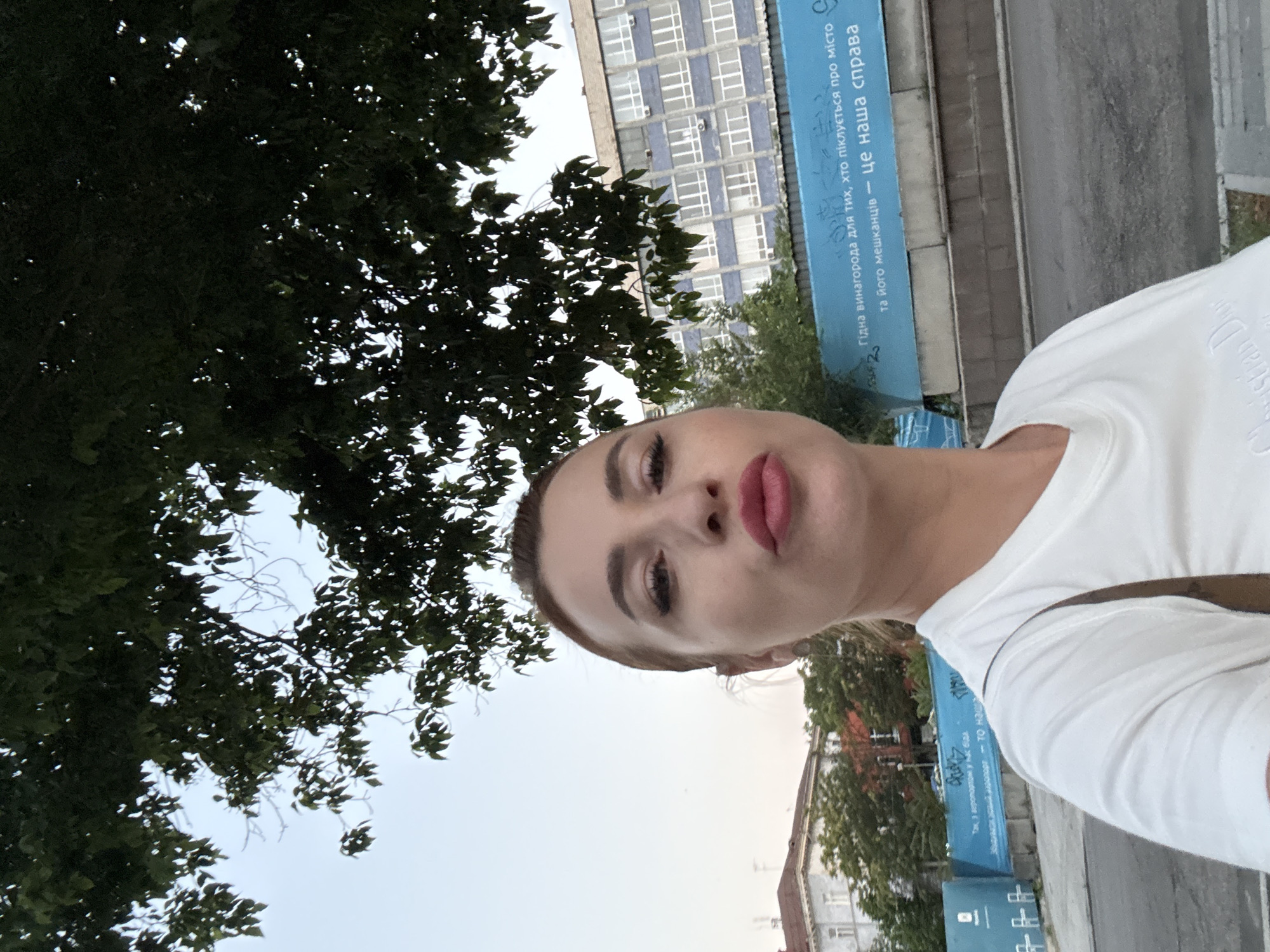 A woman in a white t-shirt takes a selfie outdoors in Dnipro, Ukraine. She has blonde hair pulled back, is wearing red lipstick, and has her lips pursed. In the background, trees with green leaves partially obscure a light blue sky. Further in the background are buildings, one with a light blue facade and many windows. A long, light blue banner with text in Ukrainian is visible, as well as a street with parked cars and a sidewalk. The banner reads "Гідна винагорода для тих, хто піклується про місто та його мешканців — це наша справа" which translates to "A worthy reward for those who care about the city and its residents is our business."