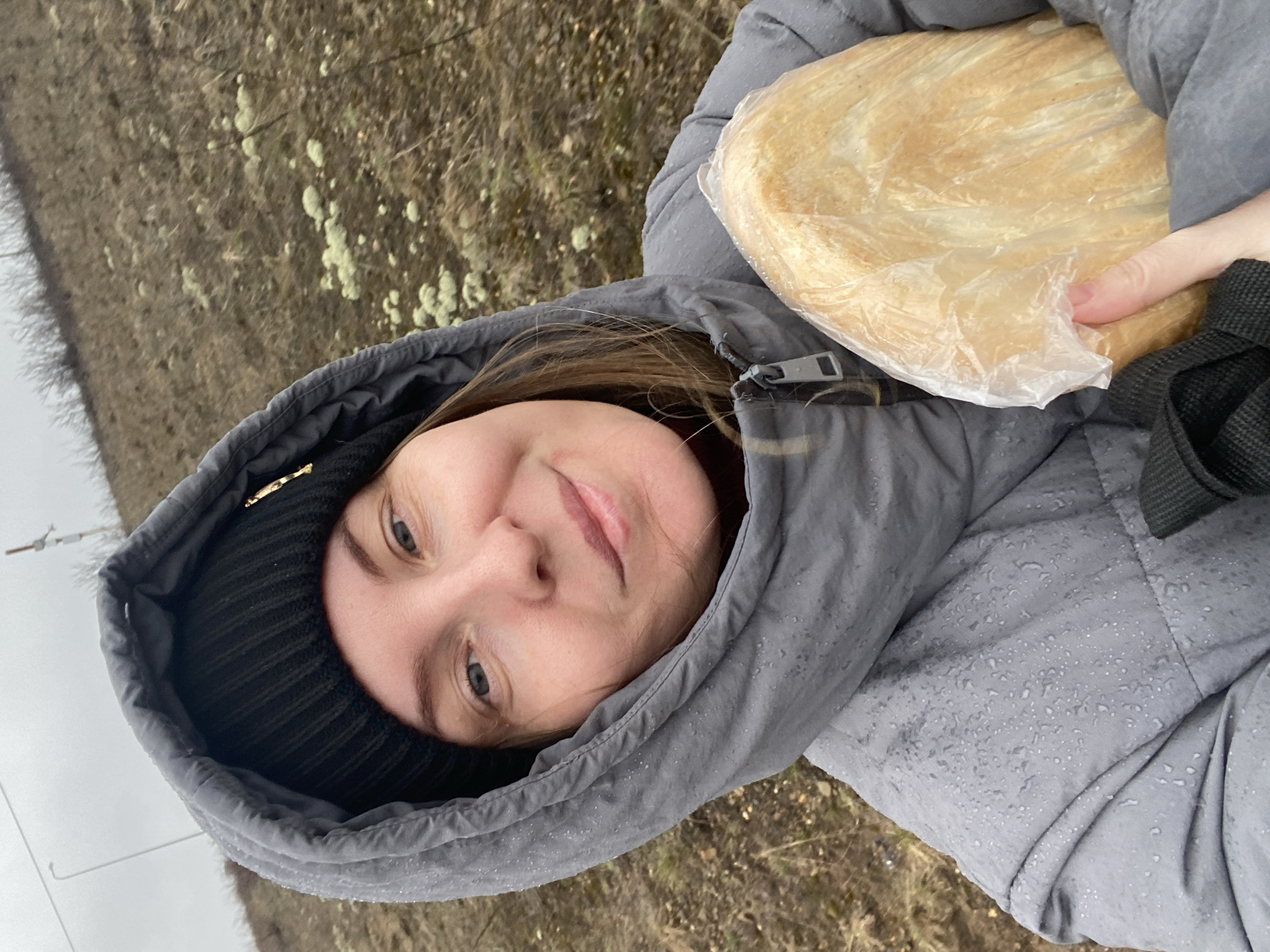 A woman wearing a black beanie and a gray hooded jacket is holding a large piece of bread wrapped in clear plastic. The jacket appears damp, with water droplets visible on the fabric. The background shows a field with sparse vegetation and some utility poles in the distance. The scene suggests a rural or semi-rural setting, with overcast weather. The location is Chaikovsky, Russia.