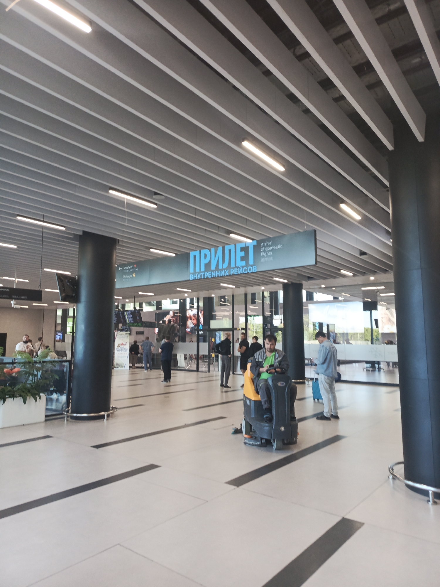 The image captures an interior view of an airport terminal. The ceiling features a modern design with parallel grey panels interspersed with white strips and illuminated by linear fluorescent lights. Two large, dark cylindrical pillars support the structure. A prominent sign in blue text reads "ПРИЛЕТ" (Arrival) followed by "Arrival of domestic flights" in English, indicating this is an arrivals hall for domestic flights.
In the foreground, a person is operating a floor cleaning machine. The machine is yellow and black and appears to be an automatic scrubber. The person is seated and wearing a green shirt. Further back, several individuals are visible. Some are standing and conversing, while others are walking. One person in the mid-ground is looking at their phone while standing next to a teal-colored suitcase. In the left mid-ground, a large plant in a white planter is visible.
The floor is tiled in a light color with dark grey rectangular inlays forming a pattern. In the background, information screens displaying flight data are visible, along with large windows that allow natural light to enter the terminal. The overall impression is of a clean and contemporary airport environment. The location is identified as the city of Ob, Russia, likely referring to Tolmachevo Airport (OVB), which serves the Novosibirsk agglomeration, including the city of Ob.