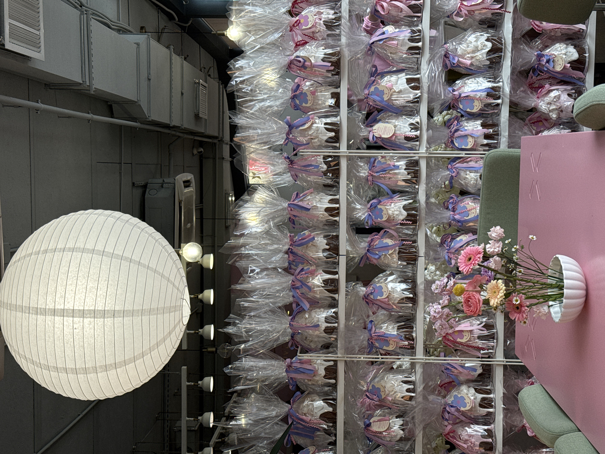 The image shows an interior scene in Moscow, Russia. On the left, a large, round, white paper lantern hangs, lit from within. The background consists of industrial-looking grey walls and ceiling, with visible ductwork and pipes. To the right, a metal rack is filled with individually wrapped pastries, each decorated with ribbons in pastel colors. The pastries appear to be displayed for sale or as decorative elements in a shop. Towards the right foreground, a pink panel with vertical lines is visible, along with a green chair. A white vase containing pink and yellow flowers is attached to the pink panel. The arrangement gives the impression of a stylish and modern interior, blending industrial elements with softer, decorative touches.