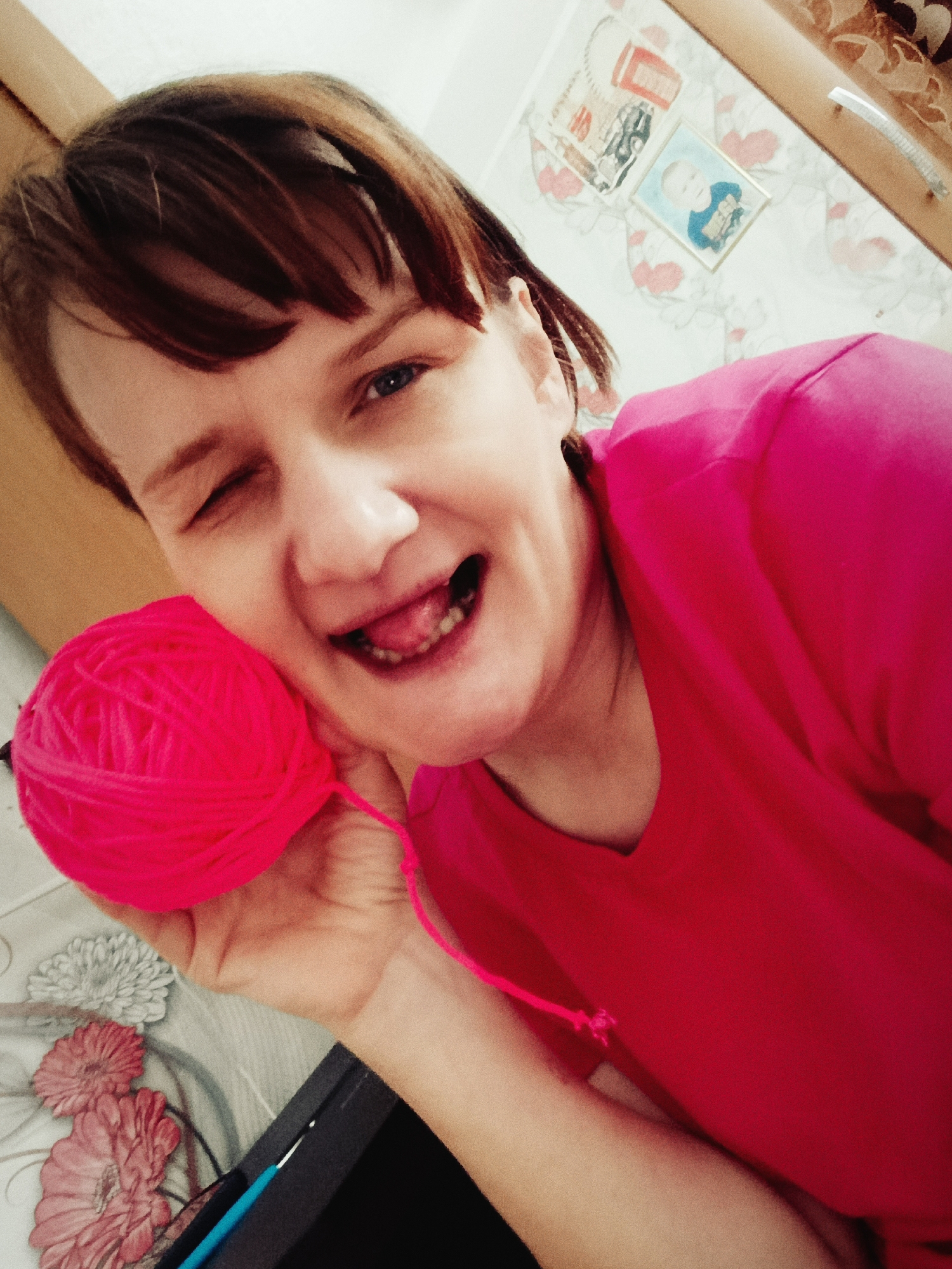 A woman with short brown hair is holding a ball of bright pink yarn in her left hand, near her face. She is wearing a pink shirt and winking with her right eye, while sticking her tongue out. The background shows a wall with floral-patterned wallpaper. There are two framed pictures on the wall; one contains a picture of a baby. A cabinet or drawer can be seen in the background on the right. The picture appears to be taken at an angle, looking downward at the woman. A crochet hook is visible in the lower left corner. There is no confirmation that the location is Фоки, Russia.