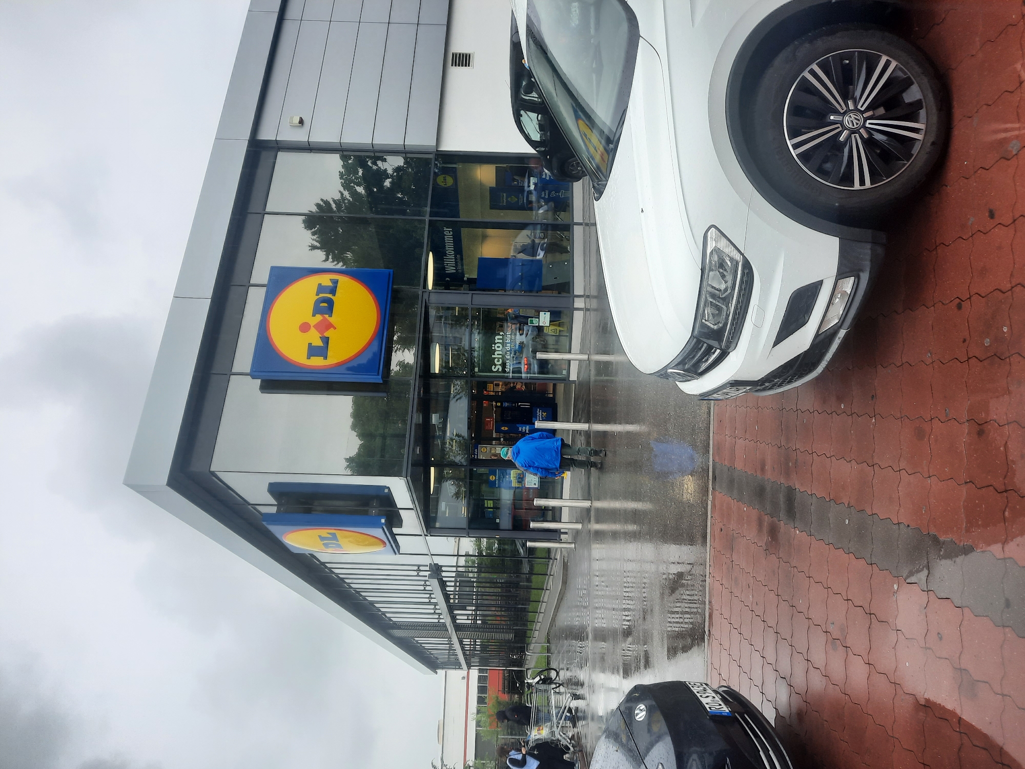 The image depicts the exterior of a Lidl supermarket in Rosenheim, Germany. The building features a modern facade with large glass windows and prominent Lidl signage, including the iconic yellow and blue logo. The foreground shows a wet, red brick paved area, indicating recent rain. Two cars are partially visible: a dark-colored car on the left and a white Volkswagen Tiguan on the right. A person in a blue jacket is walking towards the entrance of the store. Other individuals and shopping carts are visible near the entrance and in the background. The sky is overcast, contributing to a somber mood.