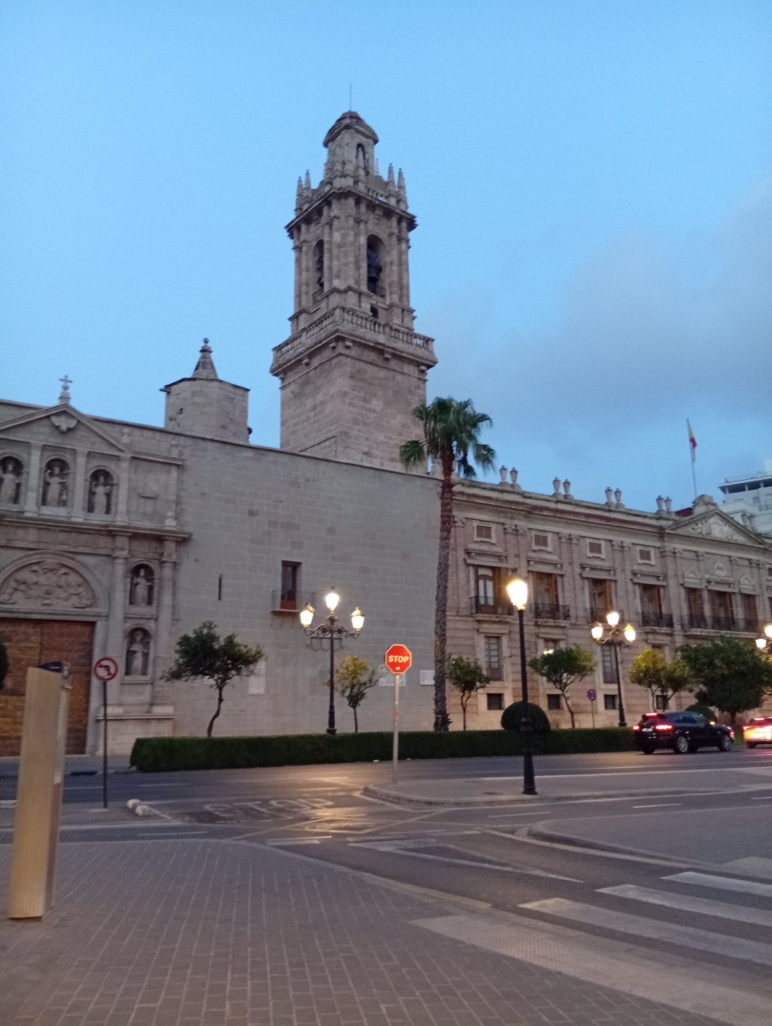 The image shows a street scene in València, Spain, featuring a large, ornate building with a bell tower, likely a church or cathedral, and an adjacent building with a classical facade. The sky is overcast and a twilight blue. Several streetlights are illuminated, casting a warm glow on the paved street and pedestrian walkway. A black car is visible driving on the road, and a red stop sign is posted on a pole. Trees and manicured hedges line the walkway in front of the buildings. The architecture suggests a historic European city.
