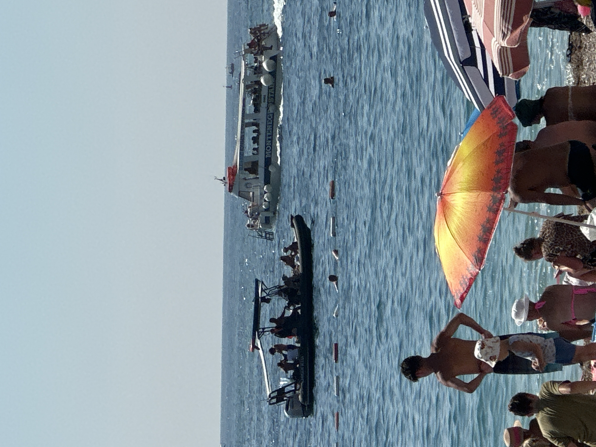 The image depicts a sunny day at a beach in Budva, Montenegro. The Adriatic Sea is visible, with several boats on the water, including a larger tour boat labeled "MONTENEGRO STAR" and a smaller rigid inflatable boat with people. In the foreground, several beachgoers are seen relaxing on the pebble beach under umbrellas. A man stands near the water, while a child in a hat stands next to him. Other people are partially visible, some sitting and others standing. The water is calm and blue, with some buoys marking a swimming area. The sky is clear and bright blue.