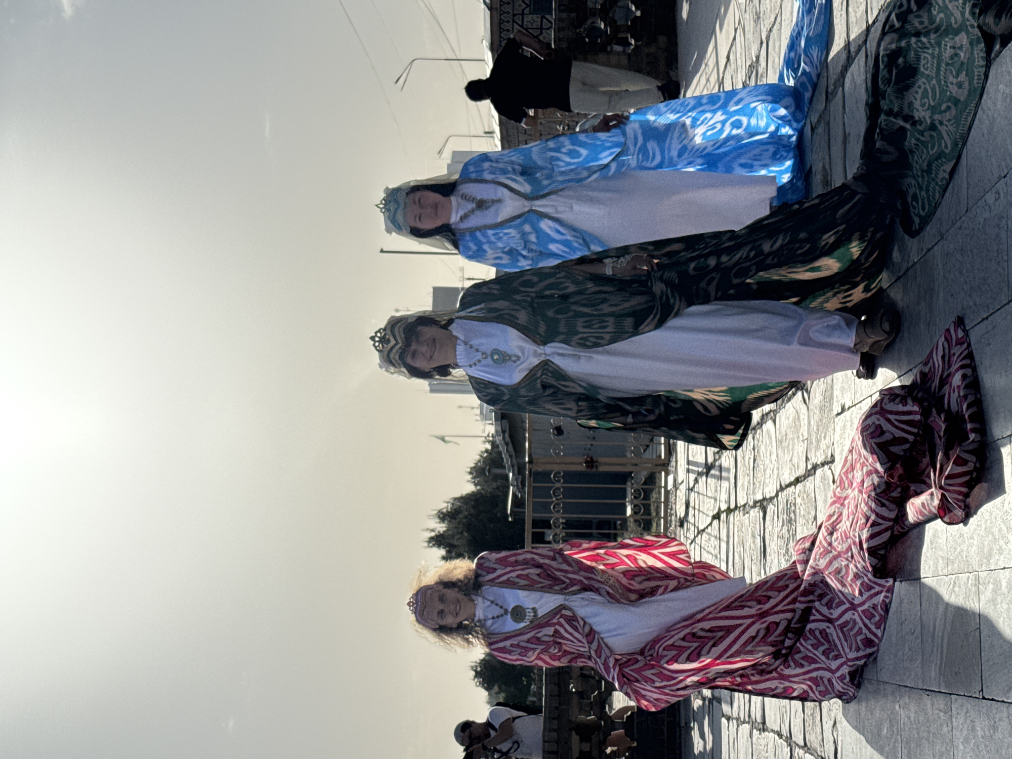 Three women in traditional, colorful ikat robes and crowns stand on a paved street in Samarkand City, Uzbekistan. Each woman wears a white dress underneath her ikat robe; one robe is red and white, another is green and white, and the third is blue and white. They face the camera. Behind them are other people, buildings, streetlights, and trees.