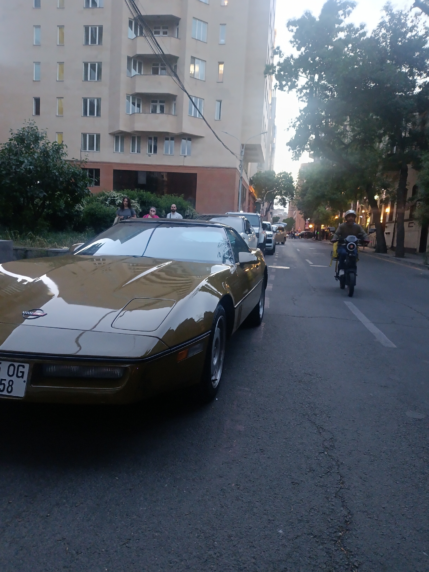 The image shows a street scene in Yerevan, Armenia. In the foreground, a gold-colored Corvette sports car is parked along the side of the road. The front of the car and part of its side are visible, along with a license plate. Behind the Corvette, other vehicles are parked, including a white pickup truck and other cars further down the street. In the middle of the road, a man on a scooter is traveling towards the camera. The man is wearing a helmet. A building stands on the left side of the street, possibly a residential building, with multiple windows and balconies. There are trees and foliage along the sidewalks, and a street lamp is visible on the right side. The environment is urban, suggesting a residential or commercial area in Yerevan.