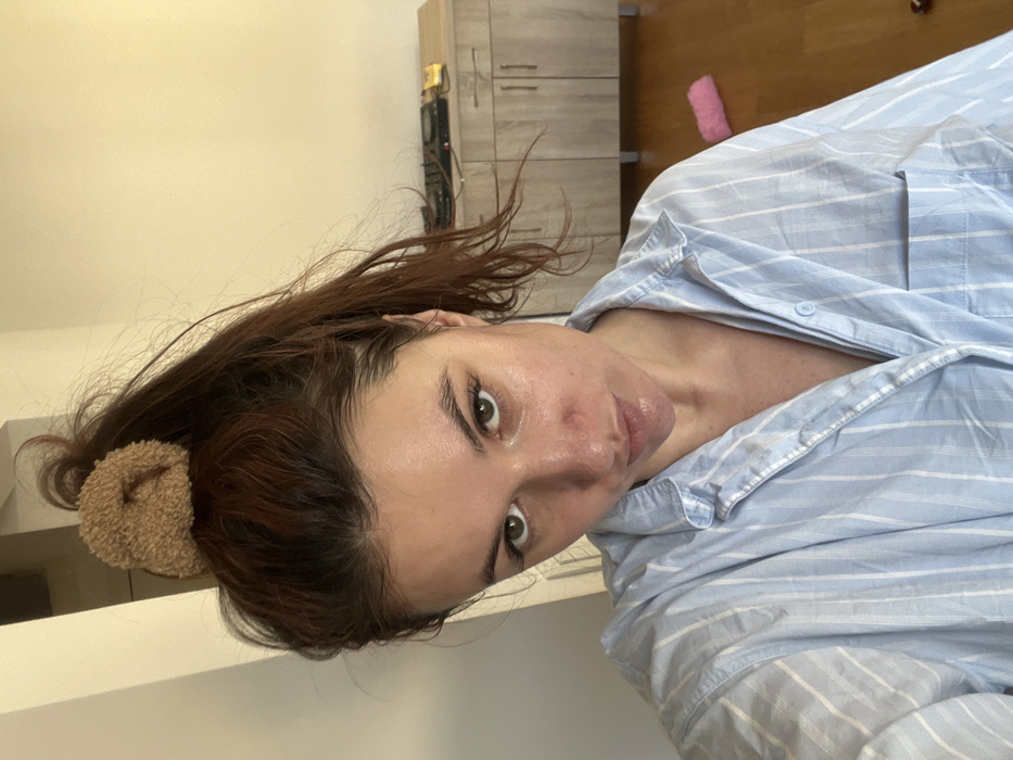 An indoor selfie captured in Budva, Montenegro, depicts a female subject from the chest up, positioned centrally. The subject has medium-length brown hair pulled into a high ponytail secured with a light brown, fuzzy scrunchie. Her eyes are green, and she looks directly into the camera with a slight pout. She is wearing a light blue and white striped button-up shirt, possibly a pajama top. The background consists of a plain, light-colored wall and a wooden floor. To the subject's right, a light-colored wooden cabinet or dresser with multiple doors is visible. On top of the cabinet, a black electronic device, possibly DJ equipment, is present, along with a small yellow object. A pink, fluffy item rests on the wooden floor to the right side of the frame. The room is evenly lit, suggesting natural light.