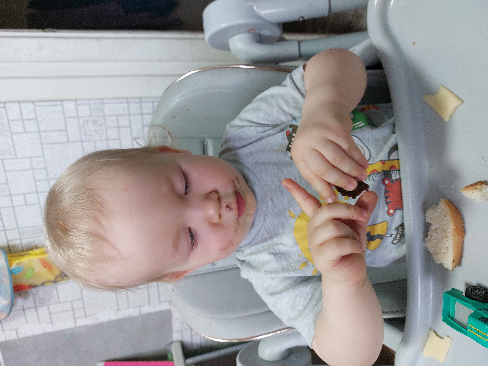 This vertical image depicts a young, light-blonde-haired child, likely a toddler, seated in a light grey high chair indoors in Chaikovsky, Russia. The child's eyes are closed, suggesting they are either asleep or resting. There are noticeable smudges of dark food, possibly chocolate, around their mouth and on their chin, indicating a recent meal.
The child is wearing a grey short-sleeved t-shirt adorned with a yellow sun design and other colorful, cartoon-like graphics, including what appears to be a road or construction scene with vehicles. Part of the word "AIRPORT" is visible on the shirt's design.
On the high chair's grey tray, several items are visible: pieces of white bread, small yellow square-shaped pieces (likely cheese), and a small, green toy car. The child's right hand is raised with two fingers extended, reminiscent of a peace sign, while the left hand is slightly curled.
The background shows a light-colored tiled floor with a grid pattern and some out-of-focus household items, indicating a home environment. The overall scene captures a calm, possibly post-meal moment, characteristic of a child's early years.